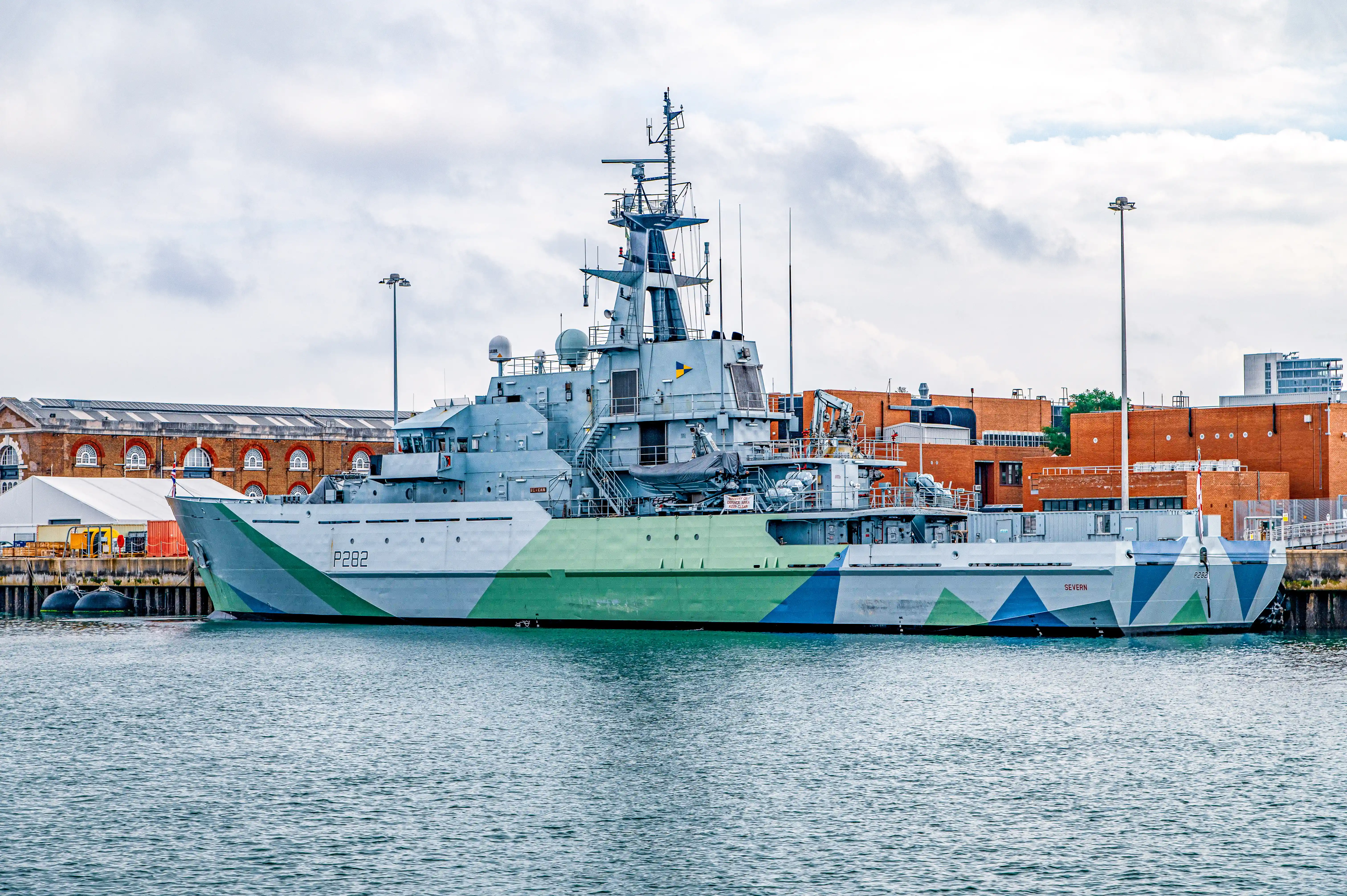 The image shows a military ship docked at a port. The ship, identified by the number P282 and the name 'Severn', features a camouflage paint scheme with green and blue geometric patterns. The ship is equipped with various antennas and radar equipment, indicating its role in surveillance or patrol missions. The background includes industrial buildings and structures, suggesting the port is in an urban or industrial area. The sky is overcast, contributing to a somewhat muted and serious atmosphere.