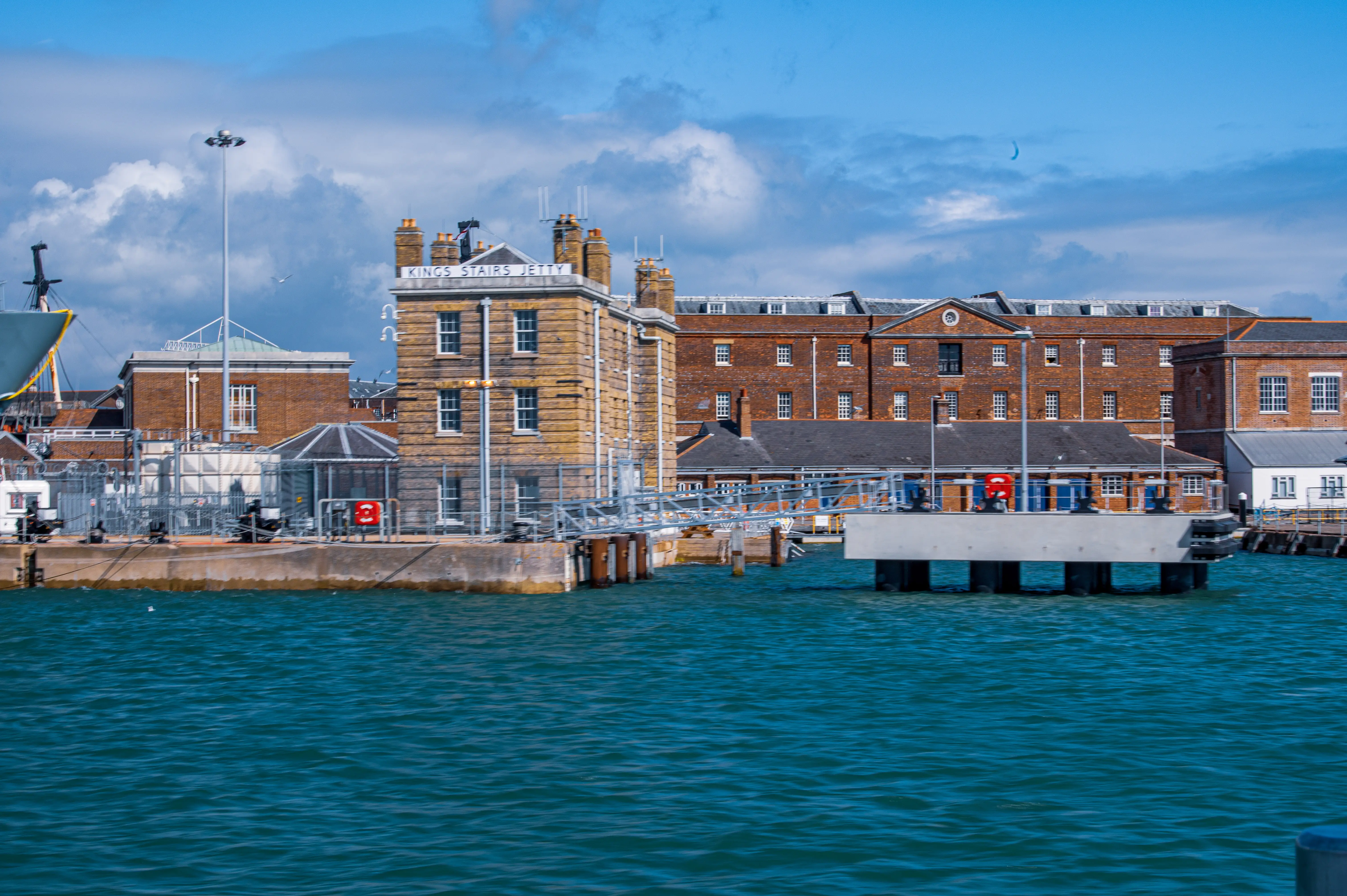 The image depicts a waterfront scene featuring a large brick building labeled 'Kings Stairs Jetty.' The building has a historic architectural style with multiple chimneys and large windows. In front of the building, there is a pier with various structures, including a small building with a pointed roof and a ramp leading down to the water. The water is a vibrant blue-green, and the sky above is partly cloudy. The overall scene suggests a maritime or port setting.