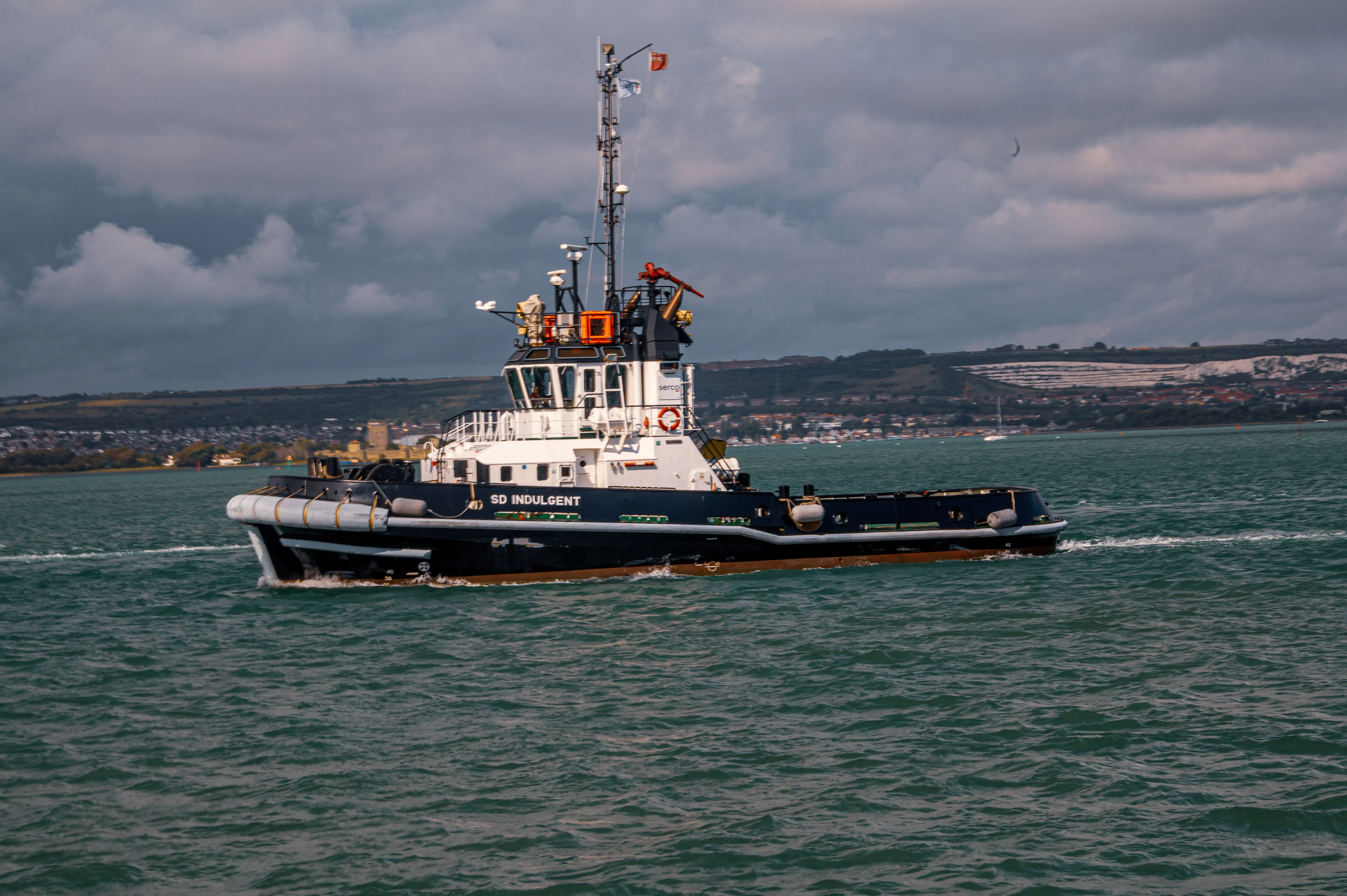 The image depicts a tugboat named 'SD Indulgent' navigating through a body of water. The vessel is equipped with various maritime equipment and is painted in dark blue and white colors. The background shows a coastal landscape with a town or city visible on the shore, under a cloudy sky.