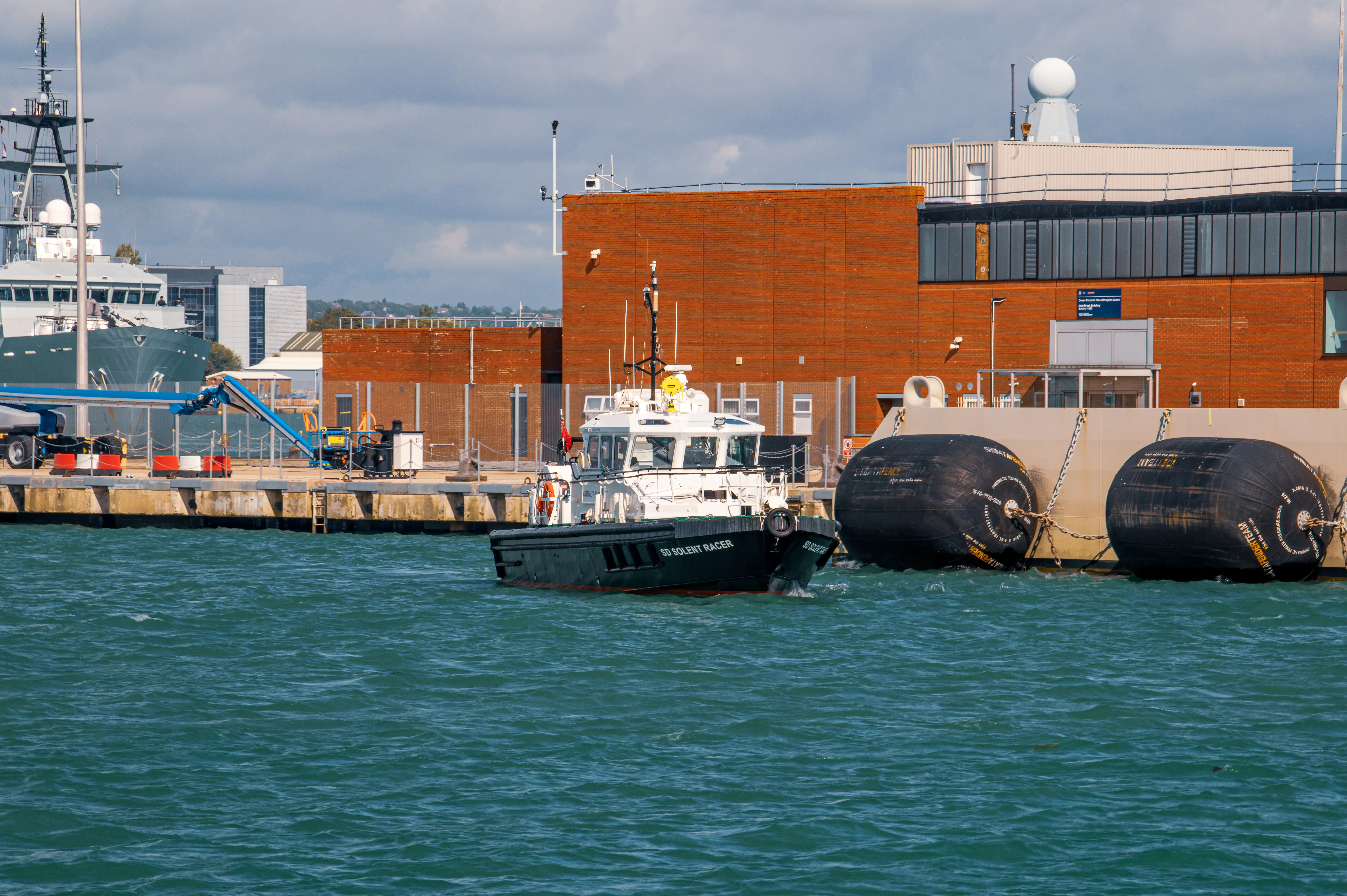 The image depicts a small boat named 'Solent Racer' navigating through a harbor. The boat is passing by large black fenders attached to a concrete pier. In the background, there is a large brick building with modern architectural features, including large windows and a white structure on the roof. Additionally, part of a larger vessel is visible to the left of the image, docked at the pier.
