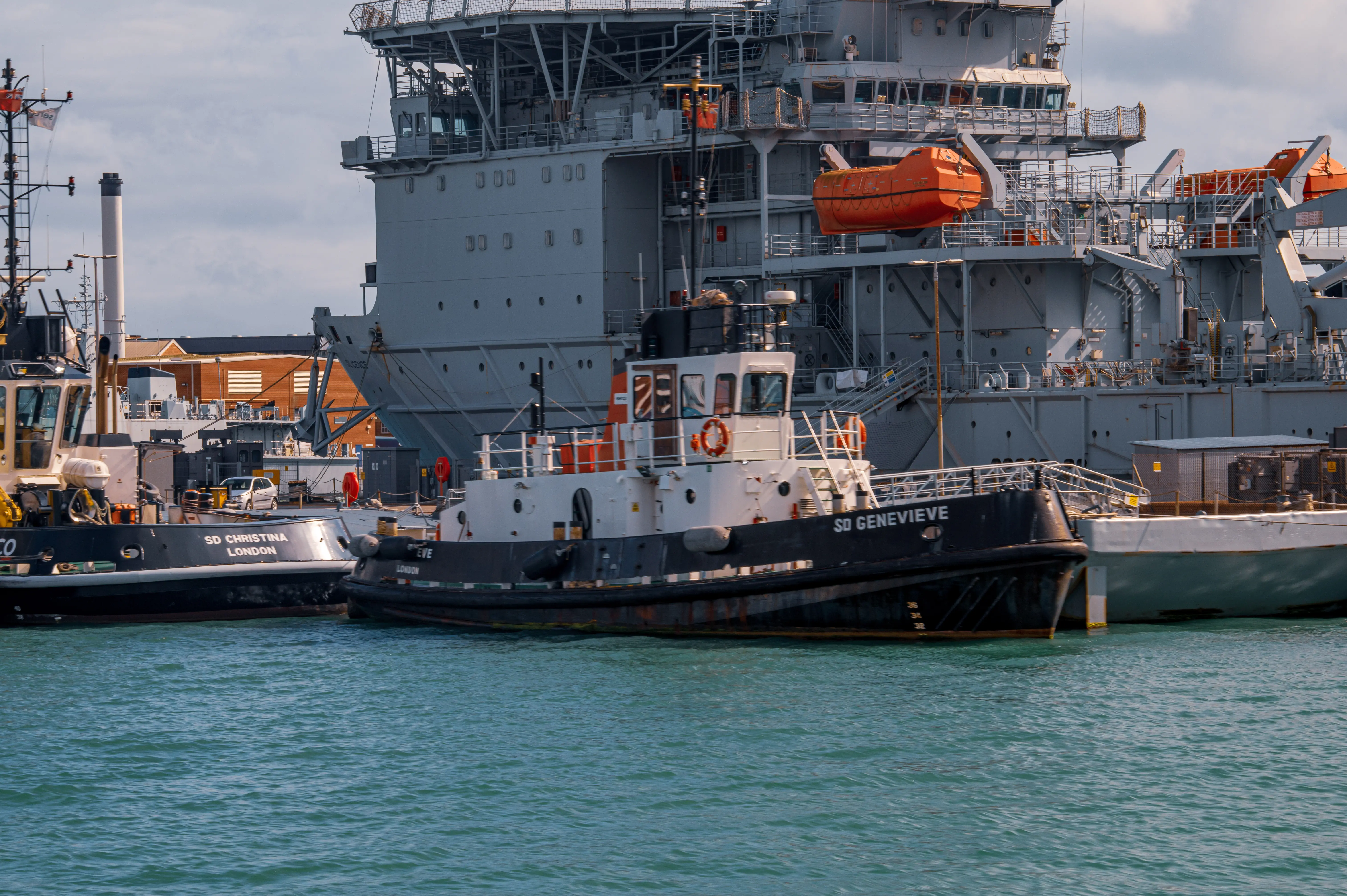 The image shows a large naval vessel docked at a port with two tugboats, named SD Genevieve and SD Christina, positioned nearby in the water. The naval vessel has various structures, equipment, and an orange lifeboat visible on its deck. The scene is set against a backdrop of industrial buildings and appears to be a busy port environment.