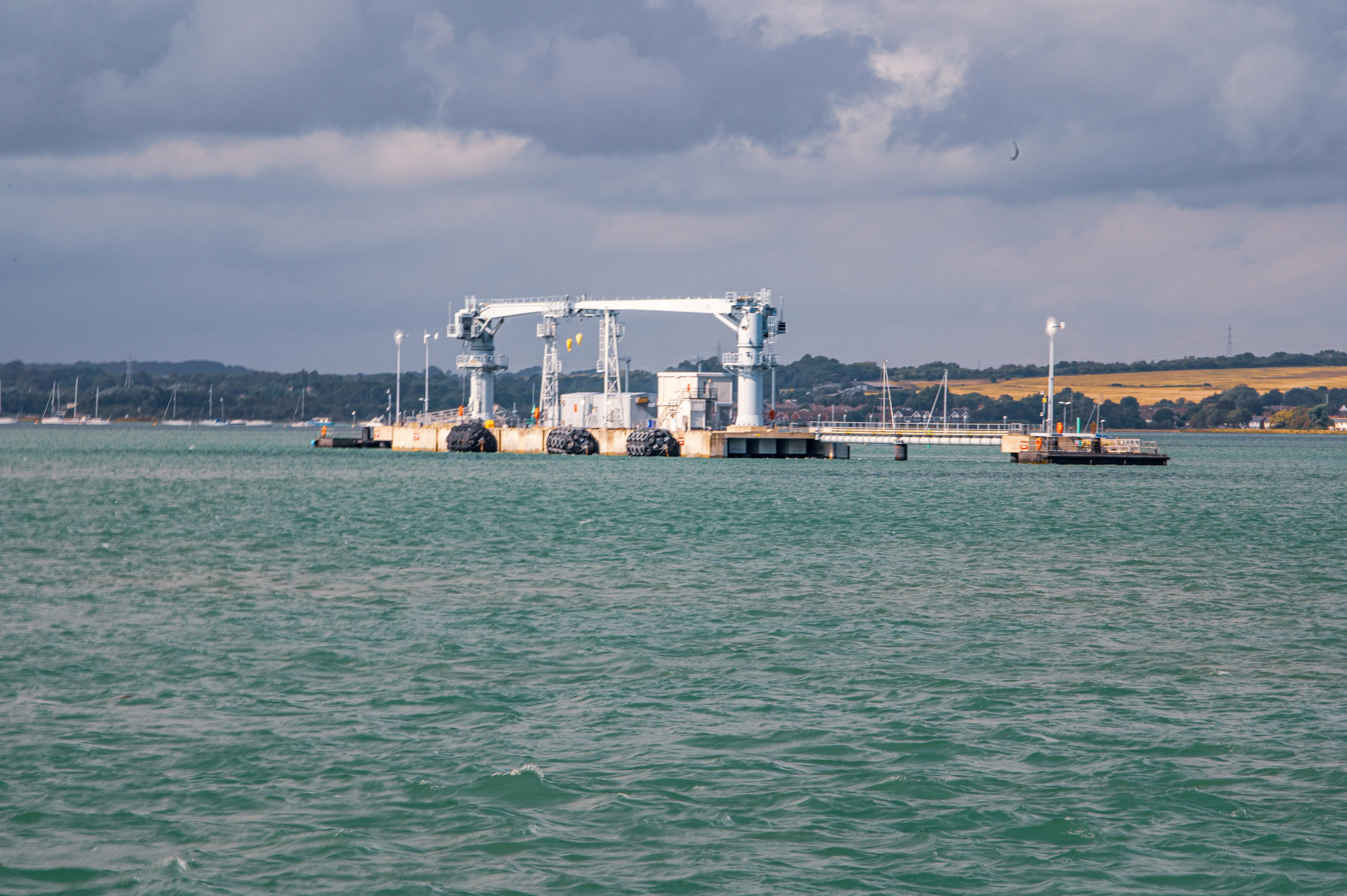 The image depicts a large industrial structure situated on a body of water, likely a port or harbor. The structure includes a significant gantry crane, which is used for heavy lifting, indicating that this area is used for loading and unloading large items, possibly shipping containers or other cargo. The surrounding area features several boats and a pier extending into the water. The background shows a coastline with trees and open fields, suggesting a mix of industrial and natural environments. The sky is overcast with clouds, and the water appears slightly choppy.