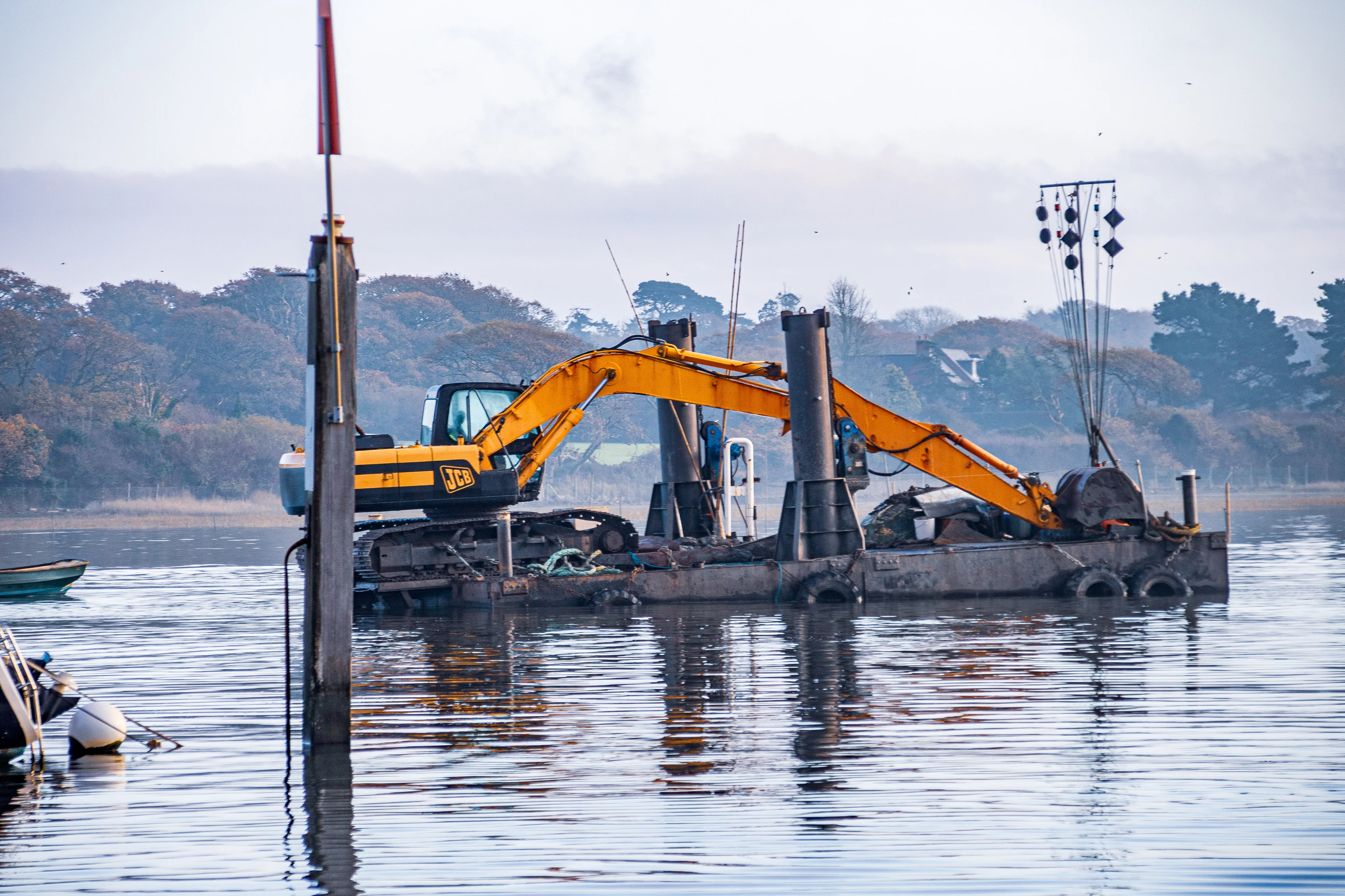 he image shows a yellow excavator on a barge in a body of water, likely engaged in construction or maintenance work. The barge is equipped with various equipment and is positioned near several vertical poles. The background features a shoreline with trees and some structures, indicating a possible riverside or lakeside setting. The scene suggests an ongoing project involving dredging, piling, or similar activities.
