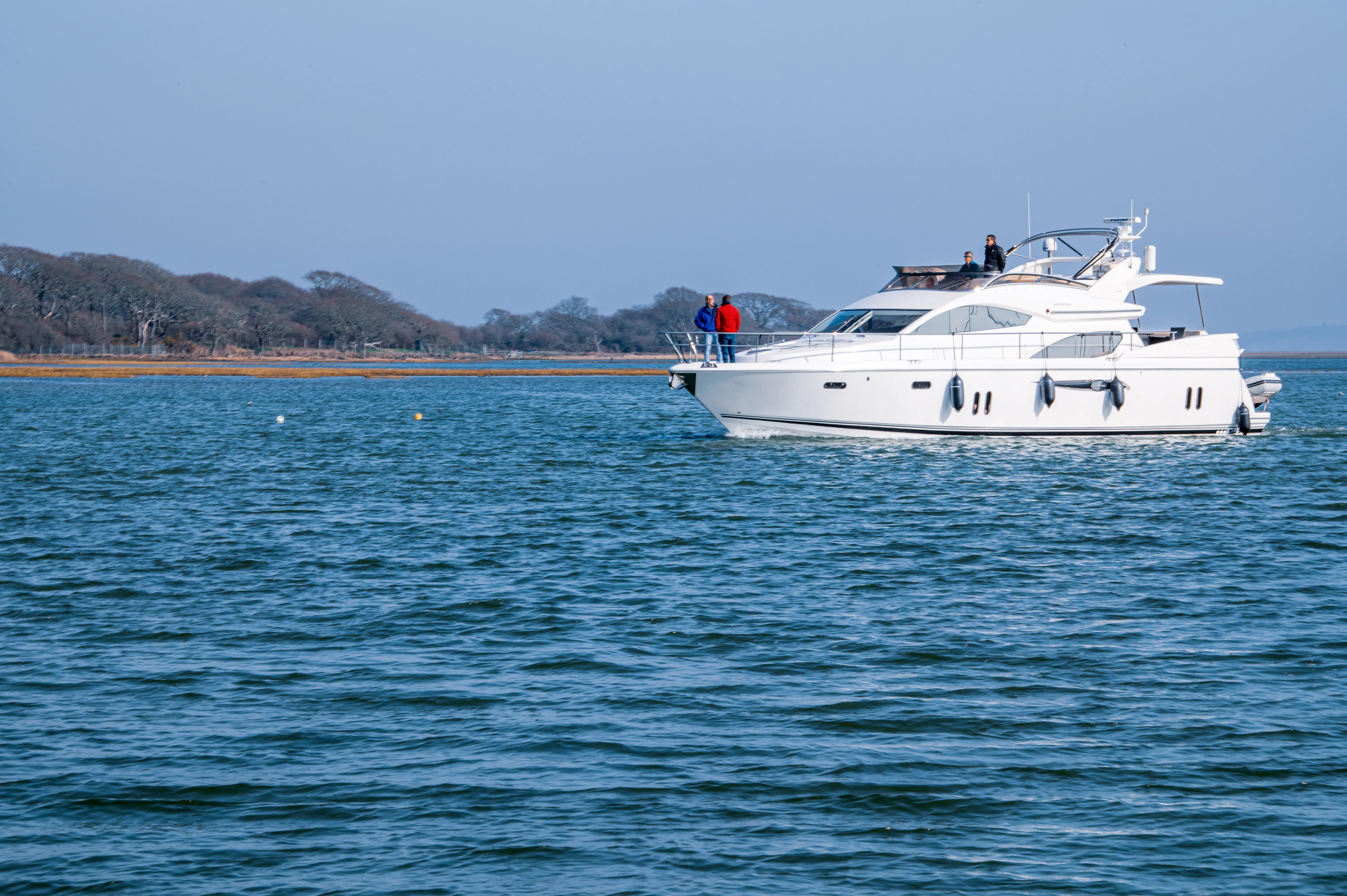 A sleek, white motor yacht glides across the blue water, carrying a few individuals. The vessel appears to be on a tranquil journey, with a treelined shore visible in the background under a clear sky.