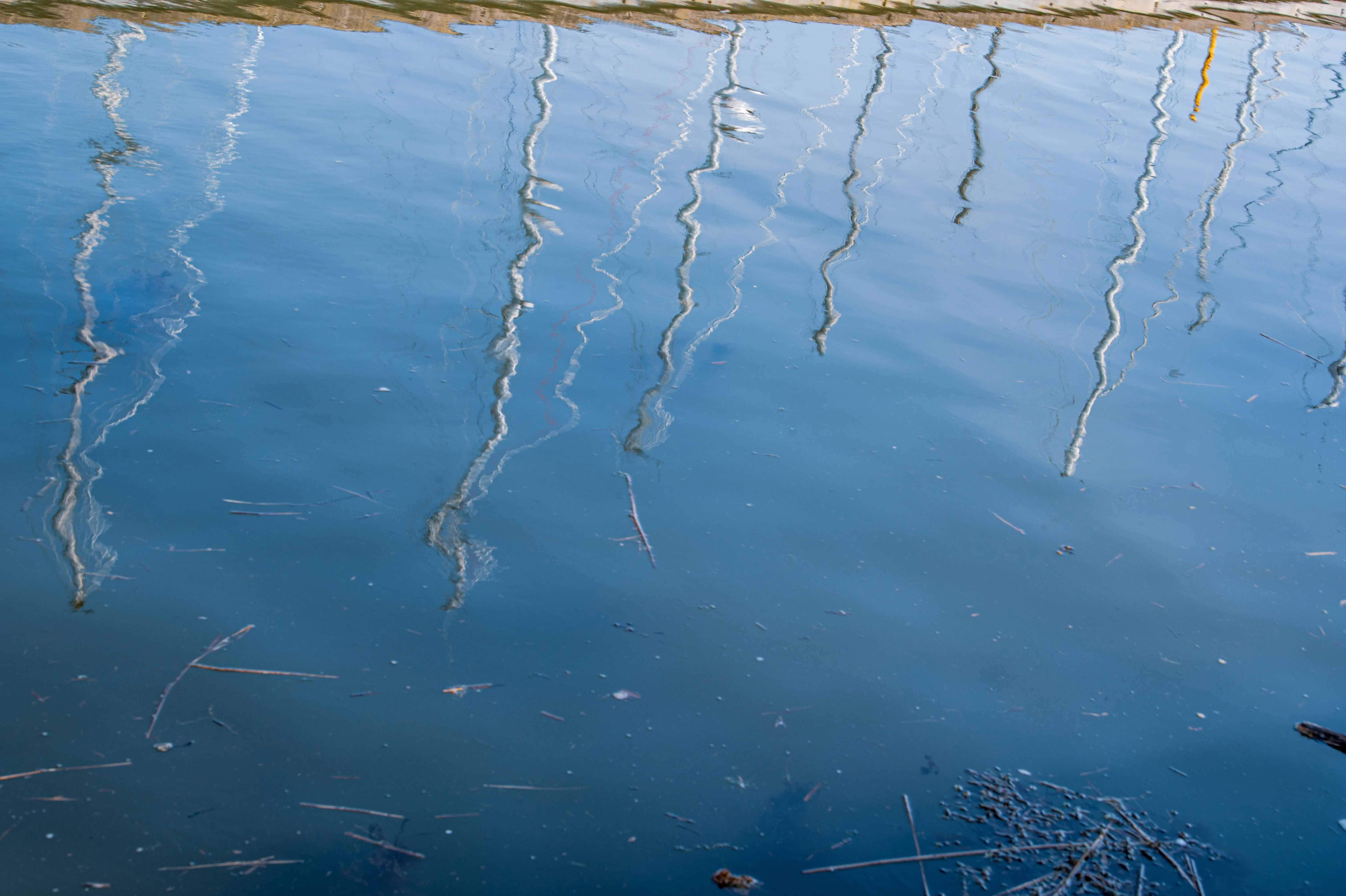 The image displays a close-up view of a body of water with reflections of what appear to be boat masts or poles extending vertically from the top. These reflections are distorted and wavy due to the movement of the water. The water itself is a deep blue, with some debris, such as small twigs and leaves, floating on the surface, particularly concentrated towards the bottom of the frame. A faint, yellowish reflection is also visible on the right side, adding a splash of color. The top edge of the image shows a hint of a beige or light brown structure, likely the edge of a dock or boat.
