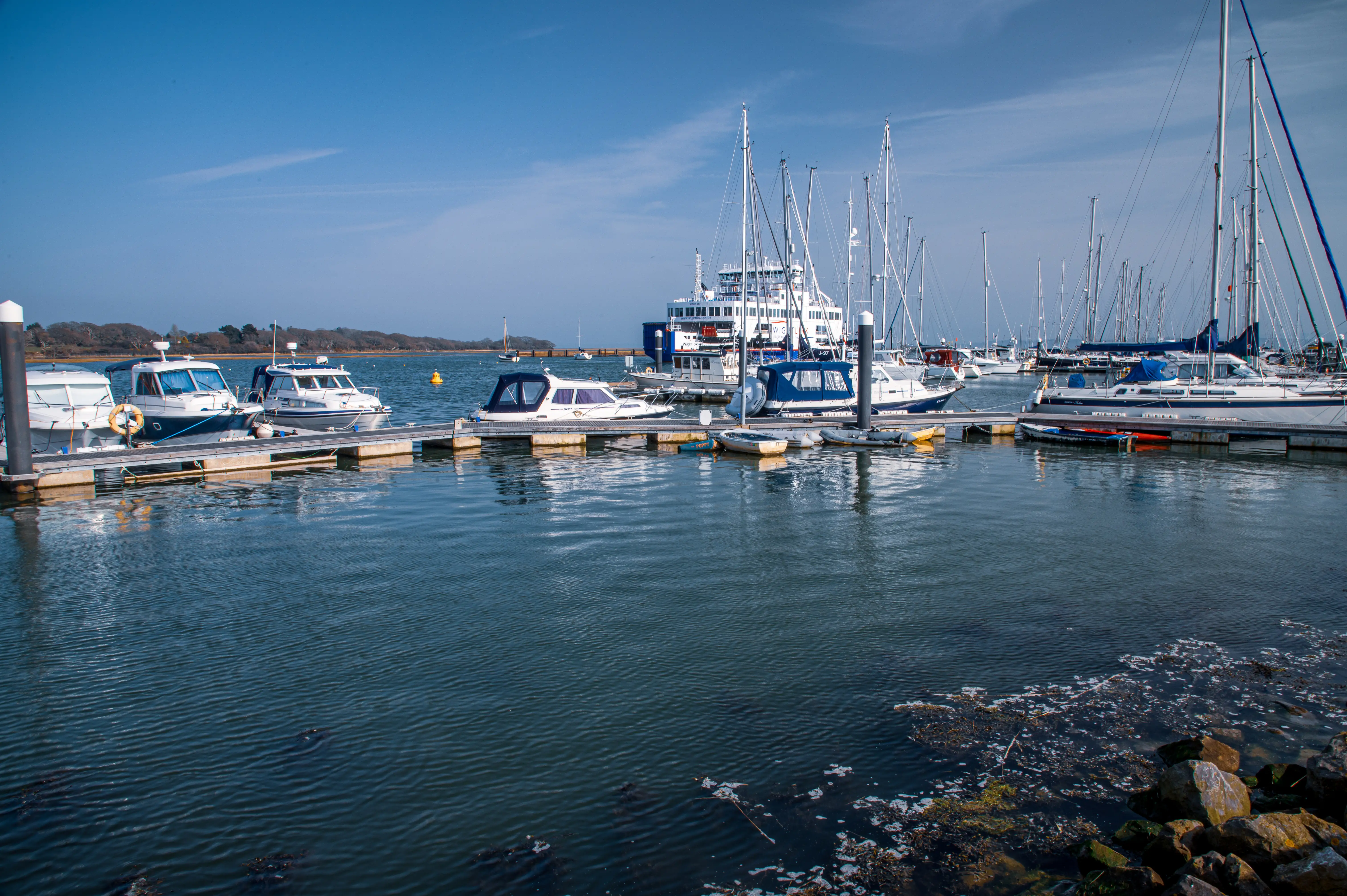 The image depicts a serene marina with various boats and yachts docked at the piers. The water is calm, reflecting the clear blue sky with a few scattered clouds. In the background, there is a large white ship with multiple decks and tall masts, likely a ferry or cruise ship. The scene is tranquil, with no visible people, suggesting a quiet day at the harbor.