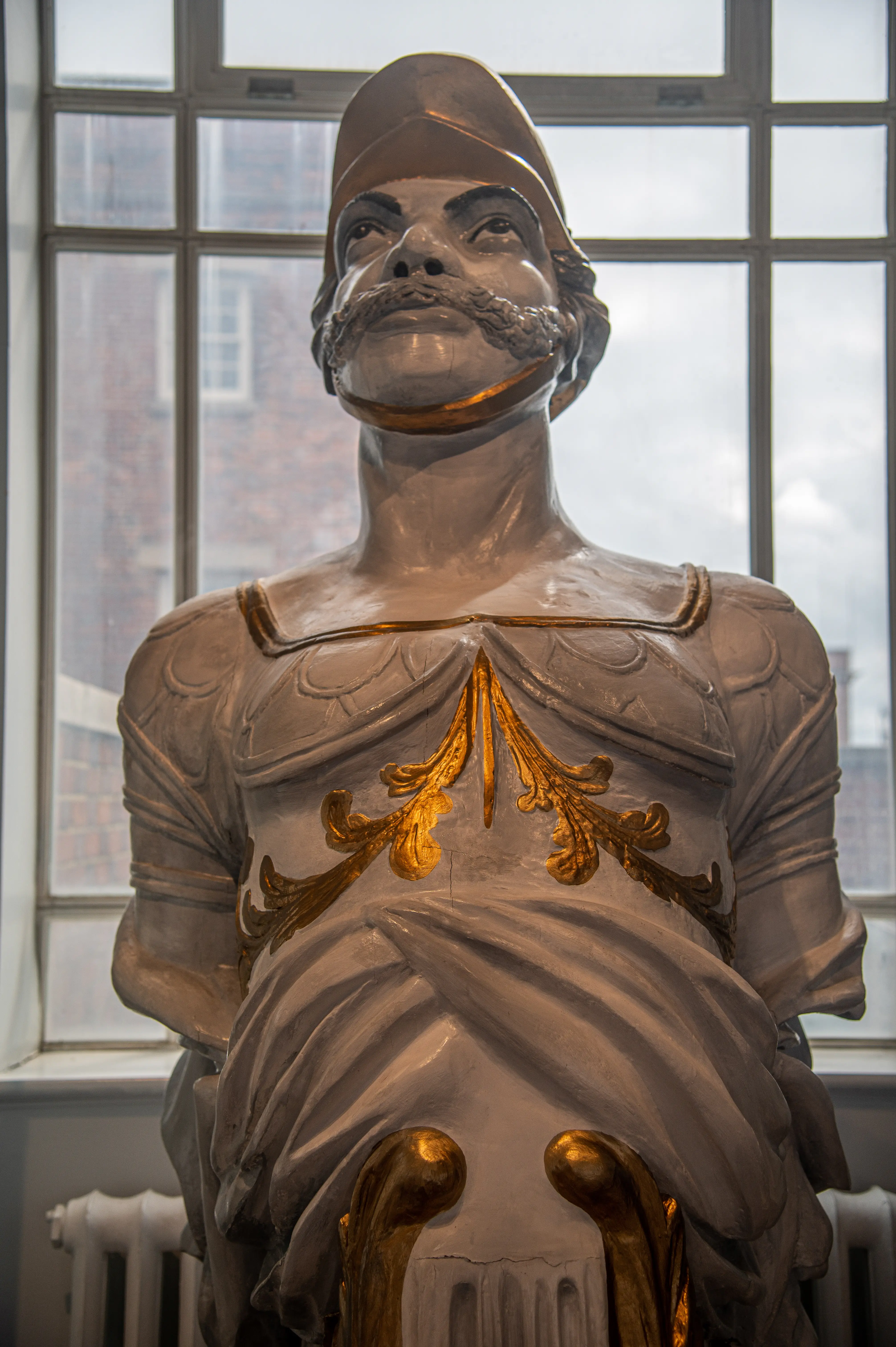 The image depicts a statue of a man wearing a helmet and a shirt with gold detailing. The statue is displayed indoors, near a window with a view of buildings outside. The man's face has distinct features, including a mustache and goatee