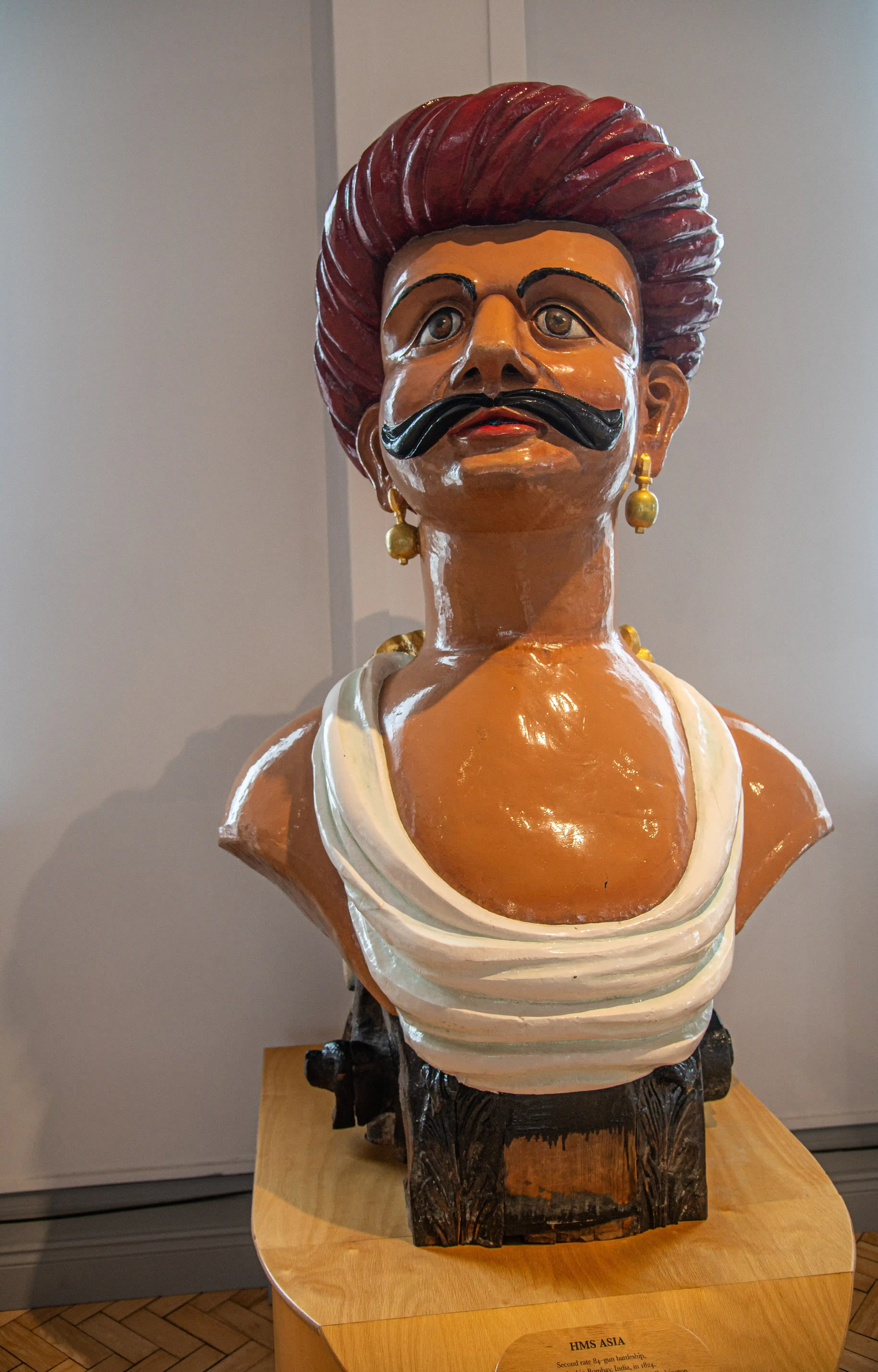 The image depicts a bust of a man with a red wig, wearing gold earrings, and adorned with a white cloth around his neck. The bust is displayed on a wooden pedestal with a plaque that reads 'HMS ASIA' and mentions a second-rate 98-gun ship launched in 1824.