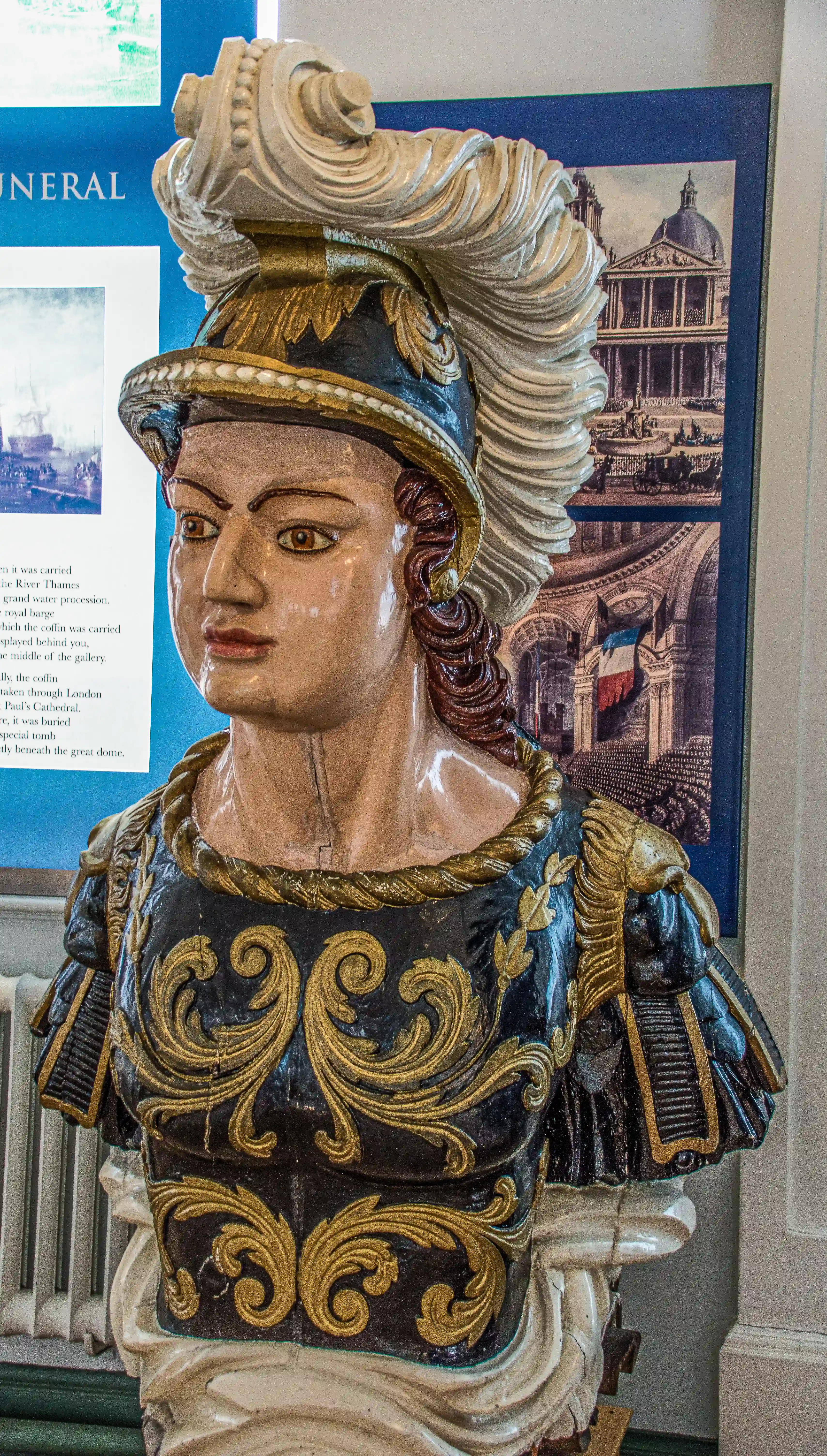 The image shows a detailed statue of a figure in elaborate armor, likely from a historical or ceremonial context. The statue features intricate designs and patterns on the armor, with a prominent helmet adorned with a large plume. The background includes historical images and text, suggesting the statue is part of an exhibit related to a significant historical event or figure.