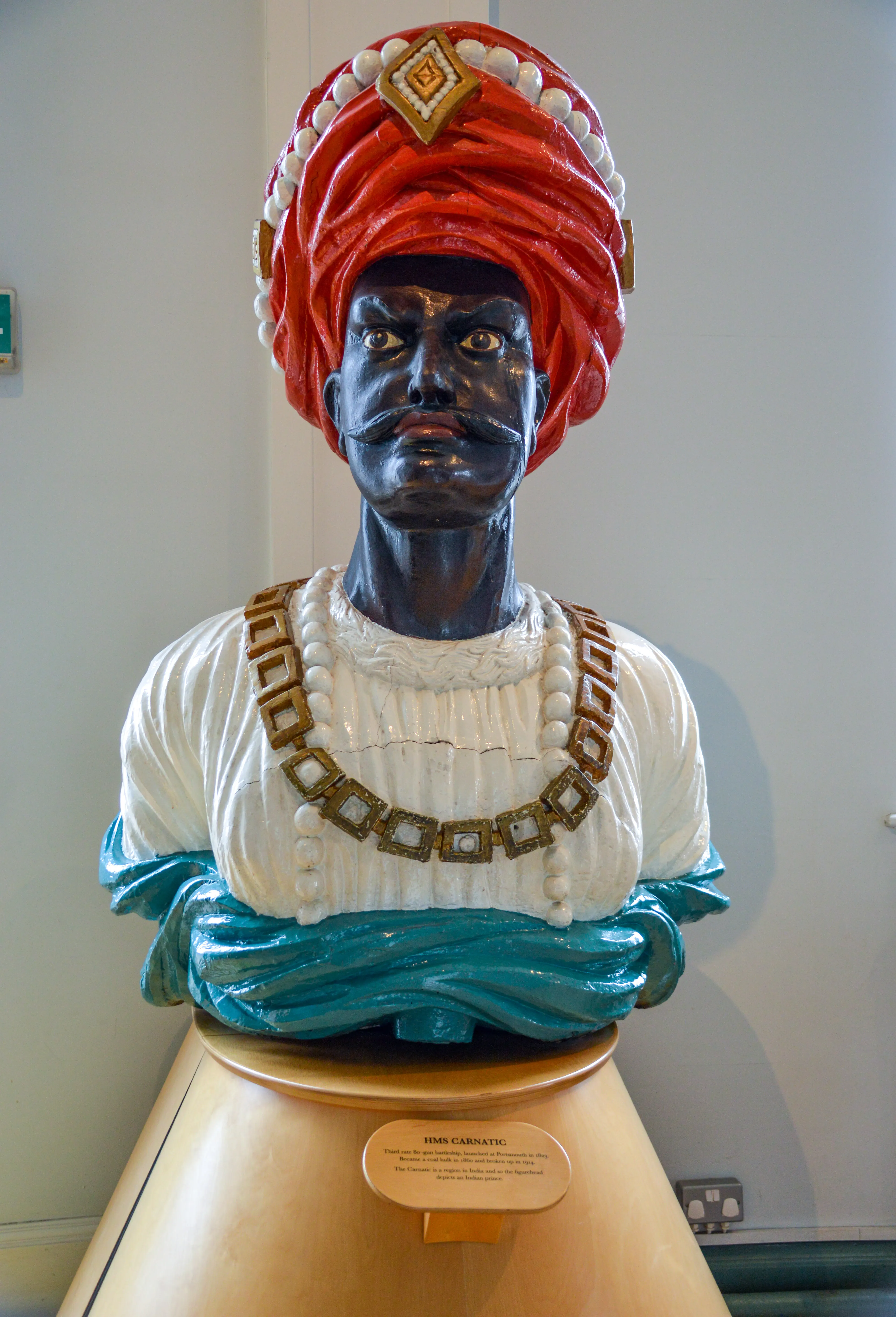 The document describes a figurehead from the HMS Carantic, a ship launched in 1822. The figurehead depicts an Indian prince, characterized by a black face with large, staring eyes, a red turban with a large jewel, a white robe adorned with a gold chain, and a blue sash. The ship was used to transport convicts to Australia and was later wrecked in 1834.