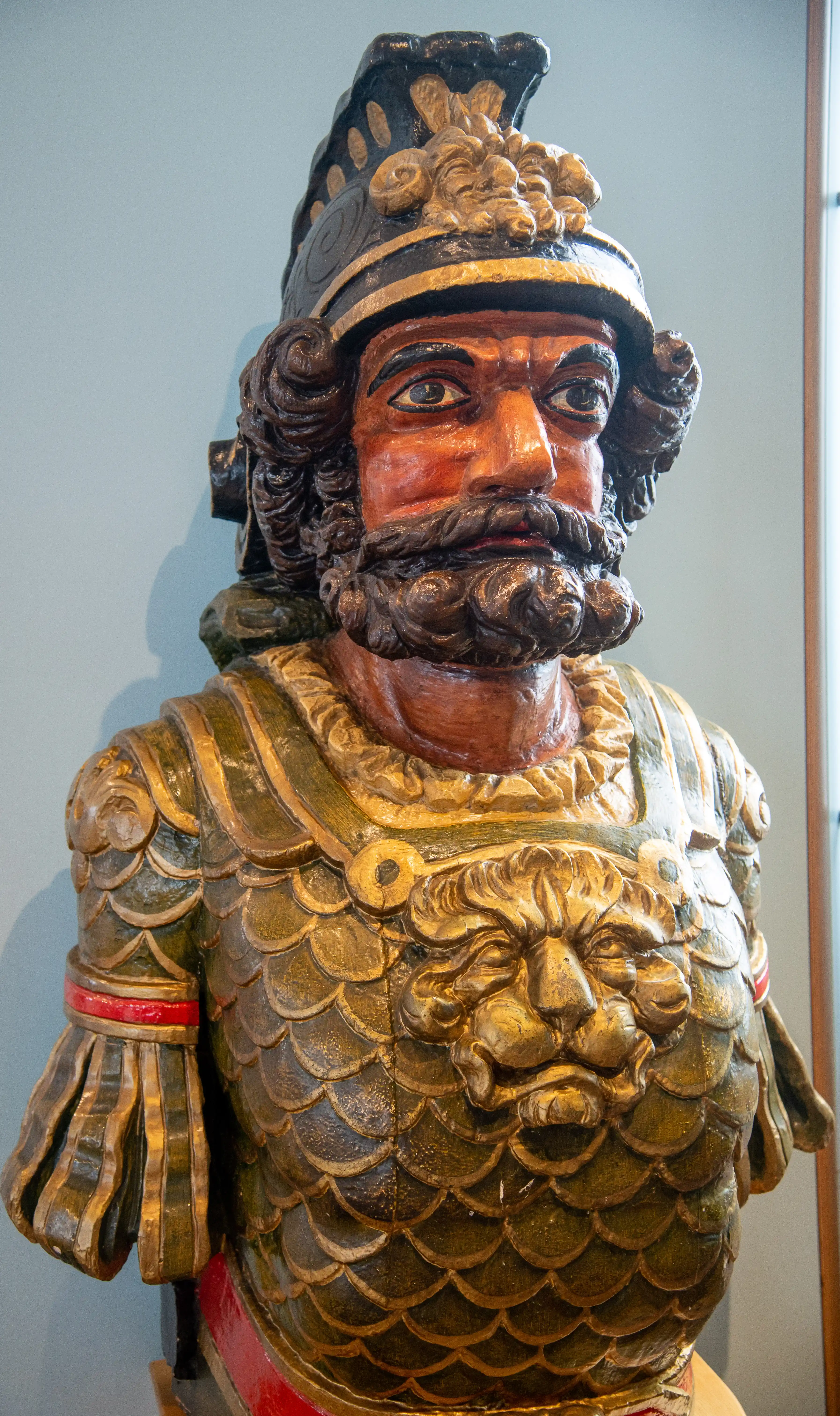 The image depicts a detailed statue of a bearded figure wearing elaborate armor. The armor is designed with a scale pattern, and the figure is adorned with a helmet featuring intricate decorations. The statue appears to be made of wood and painted with rich colors, giving it a lifelike and historical appearance. The figure's intense expression and detailed craftsmanship suggest it may represent a historical or mythical warrior.