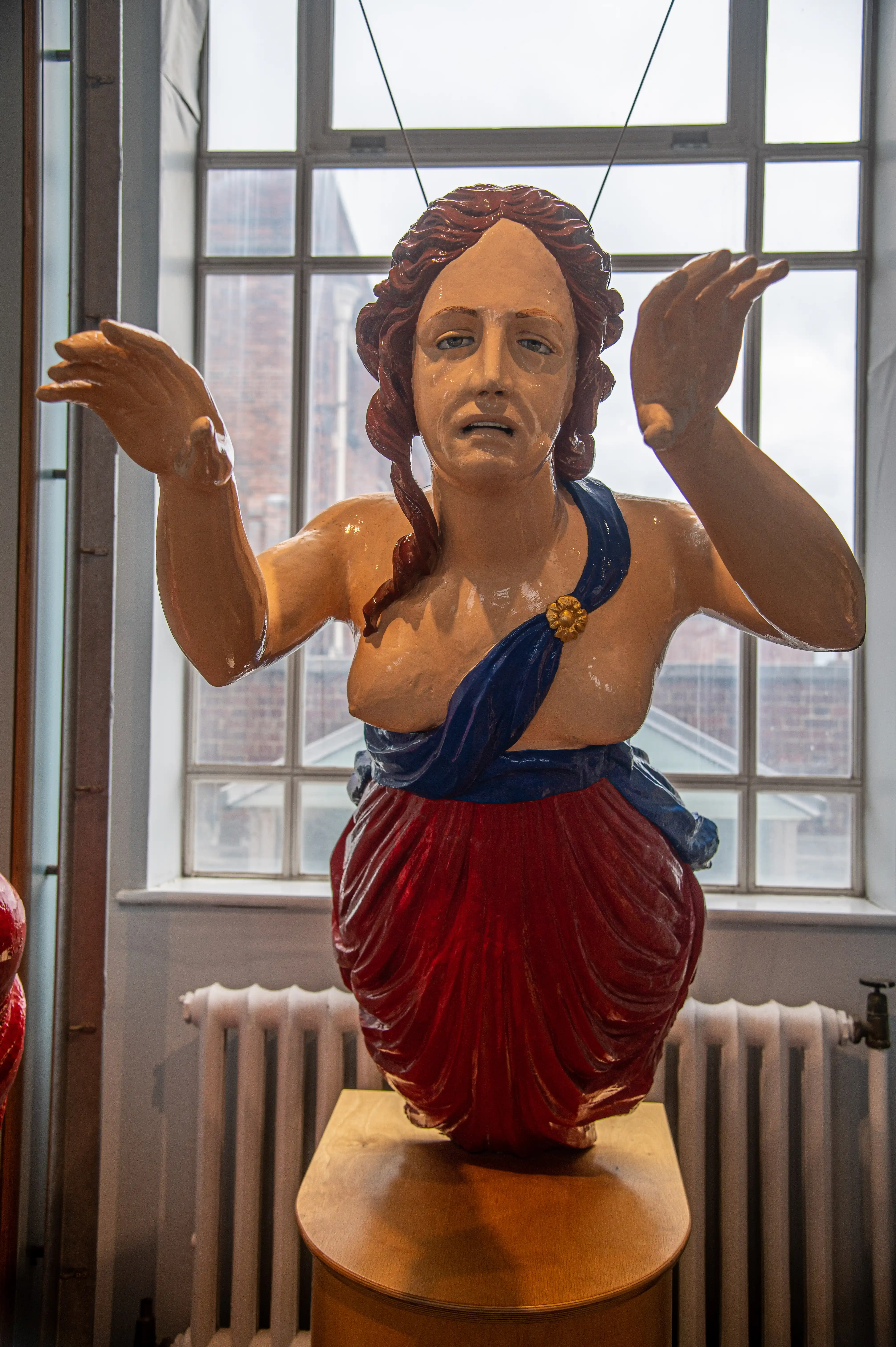 The image depicts a statue of a woman with a dramatic expression, wearing a blue and red garment, and positioned in front of a window with a view of buildings outside. The statue is placed on a wooden pedestal and is situated indoors, near a radiator.