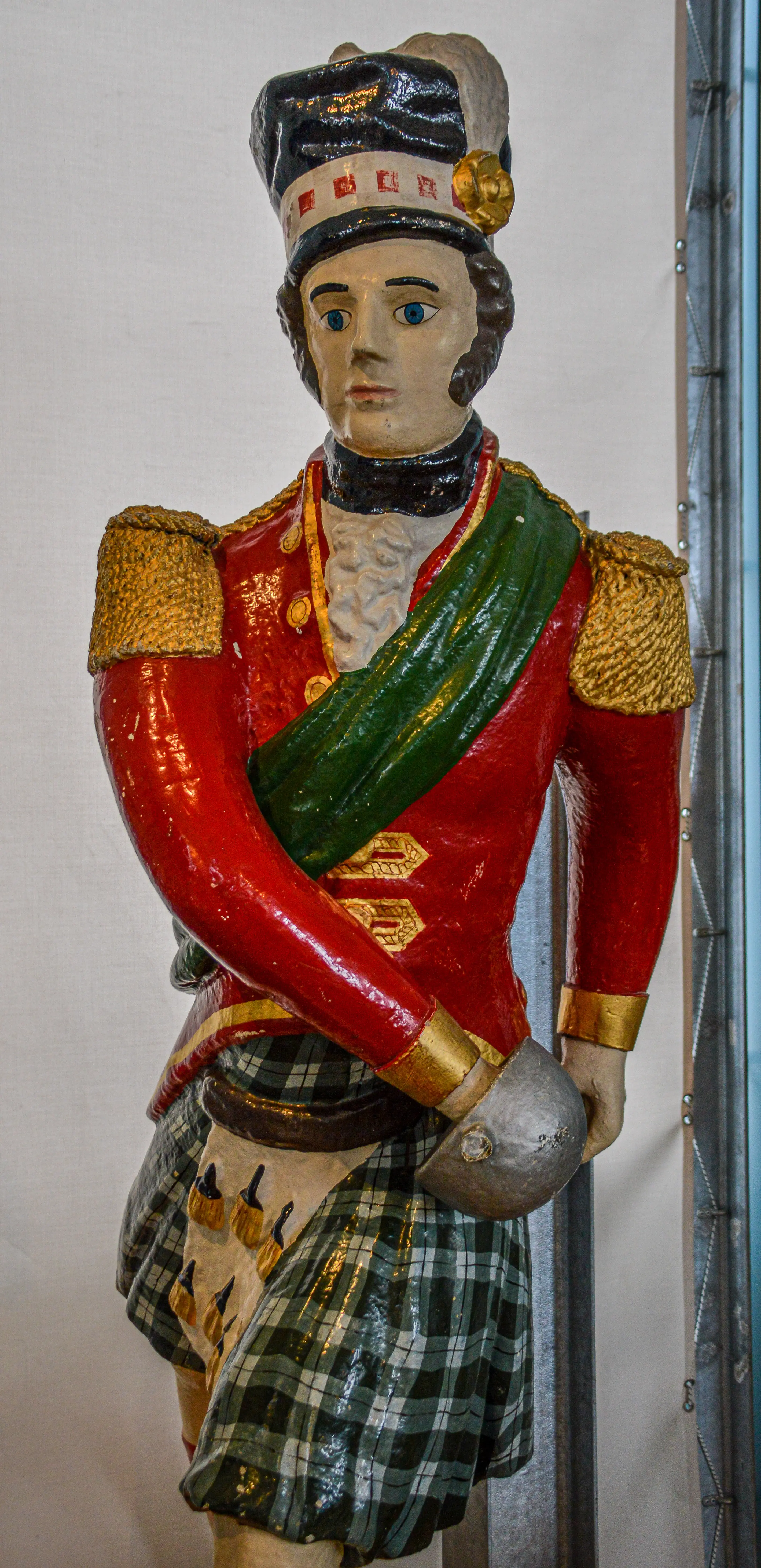 The image depicts a statue of a historical soldier dressed in traditional Highland military attire. The figure is adorned with a red jacket with gold accents, a green sash, a kilt with a plaid pattern, and a black hat with a red and gold band. The statue holds a sword in its right hand, resting it on a grey stone. The figure's face is painted with detailed features, including blue eyes and a serious expression. The overall appearance suggests a representation of a Scottish soldier from a bygone era.