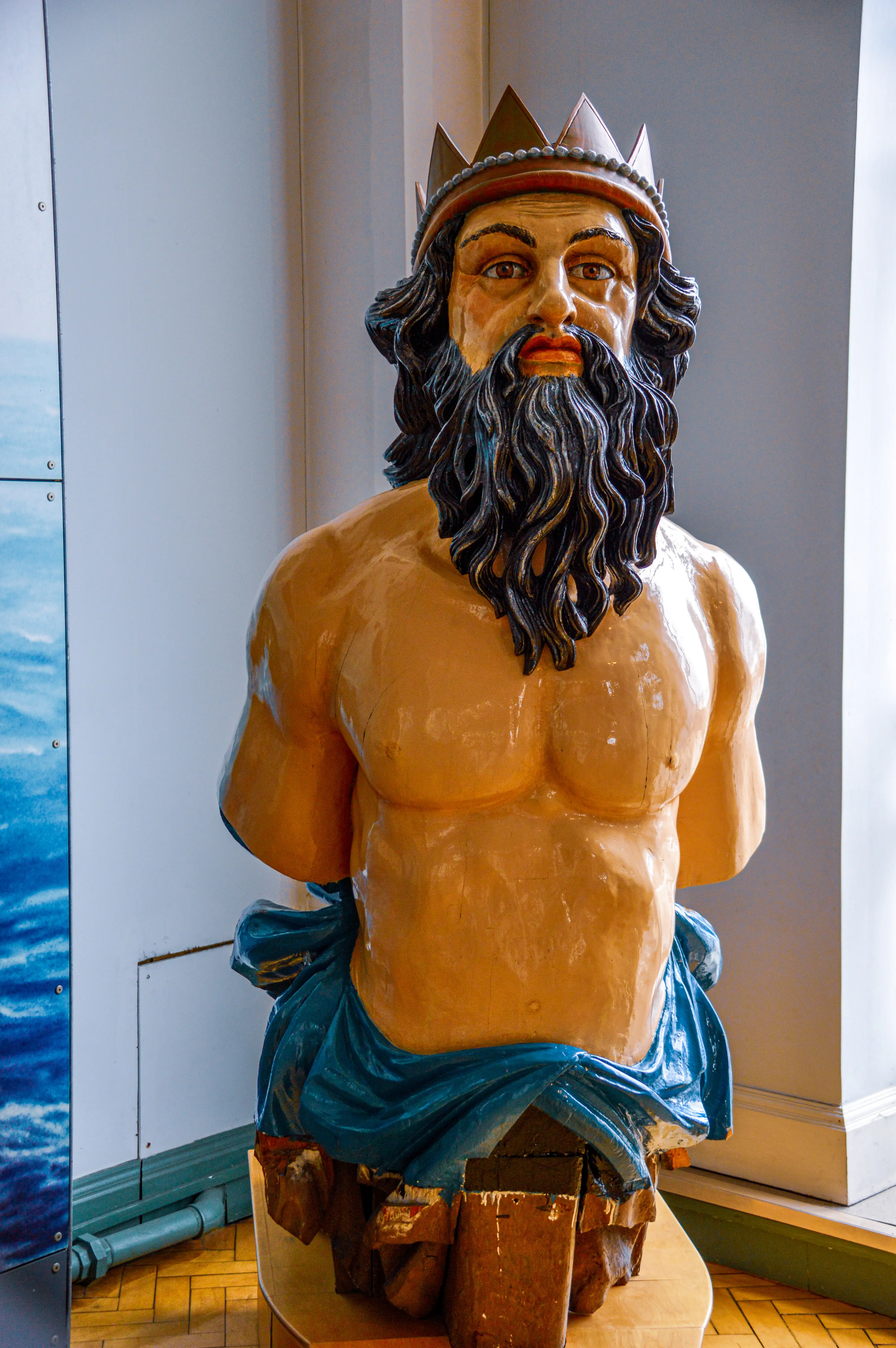 The image depicts a statue of a muscular, bearded man wearing a crown, draped in a blue cloth around his waist, seated on a wooden structure. The statue appears to be indoors, with a window and some pipes visible in the background.