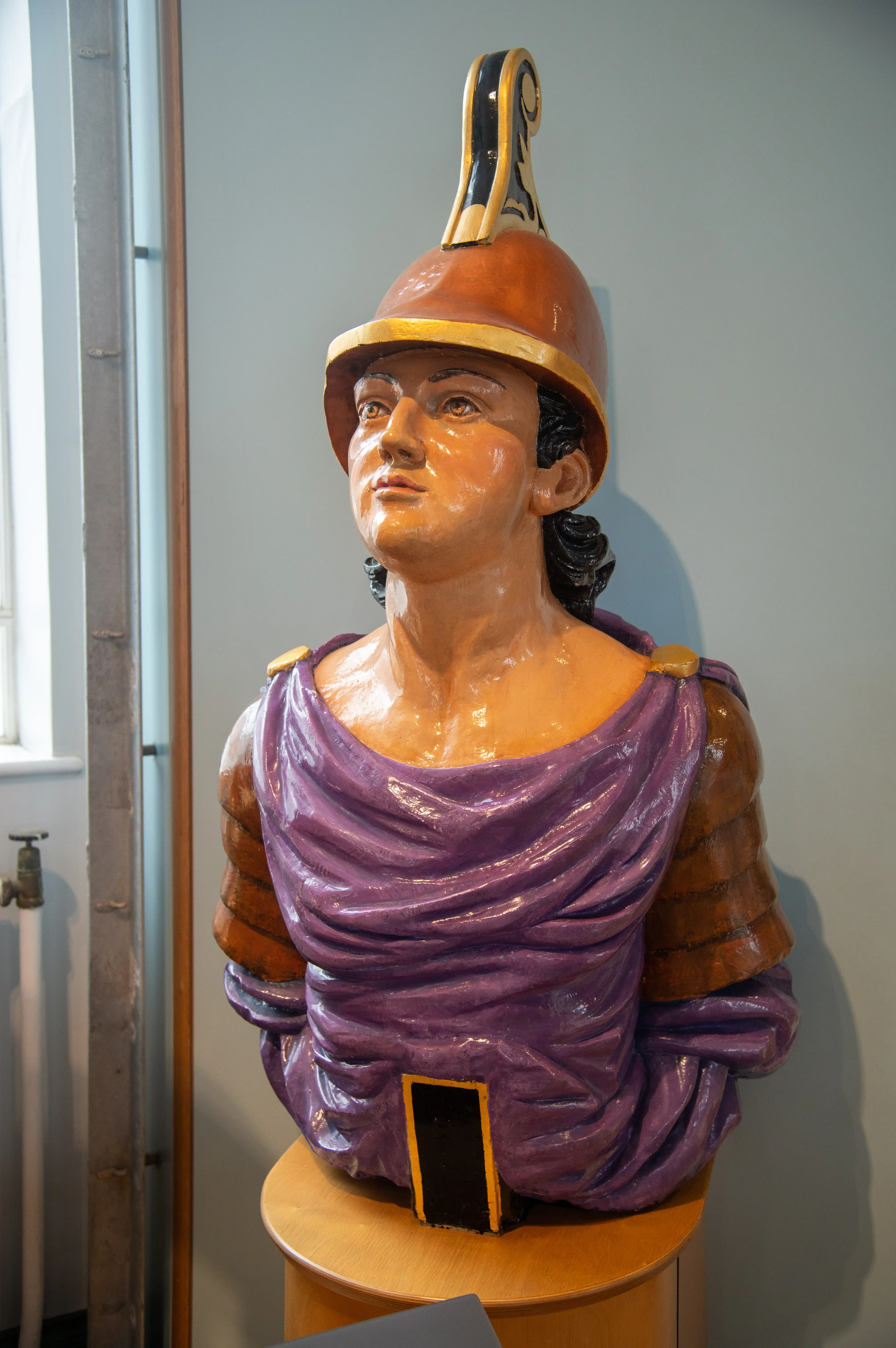 The image depicts a bust of a person wearing a purple garment and an orange helmet with a plume. The bust is placed on a wooden pedestal and is positioned indoors, possibly in a museum or gallery setting. The person's expression is serious, and the bust is detailed, showcasing the craftsmanship involved in its creation.