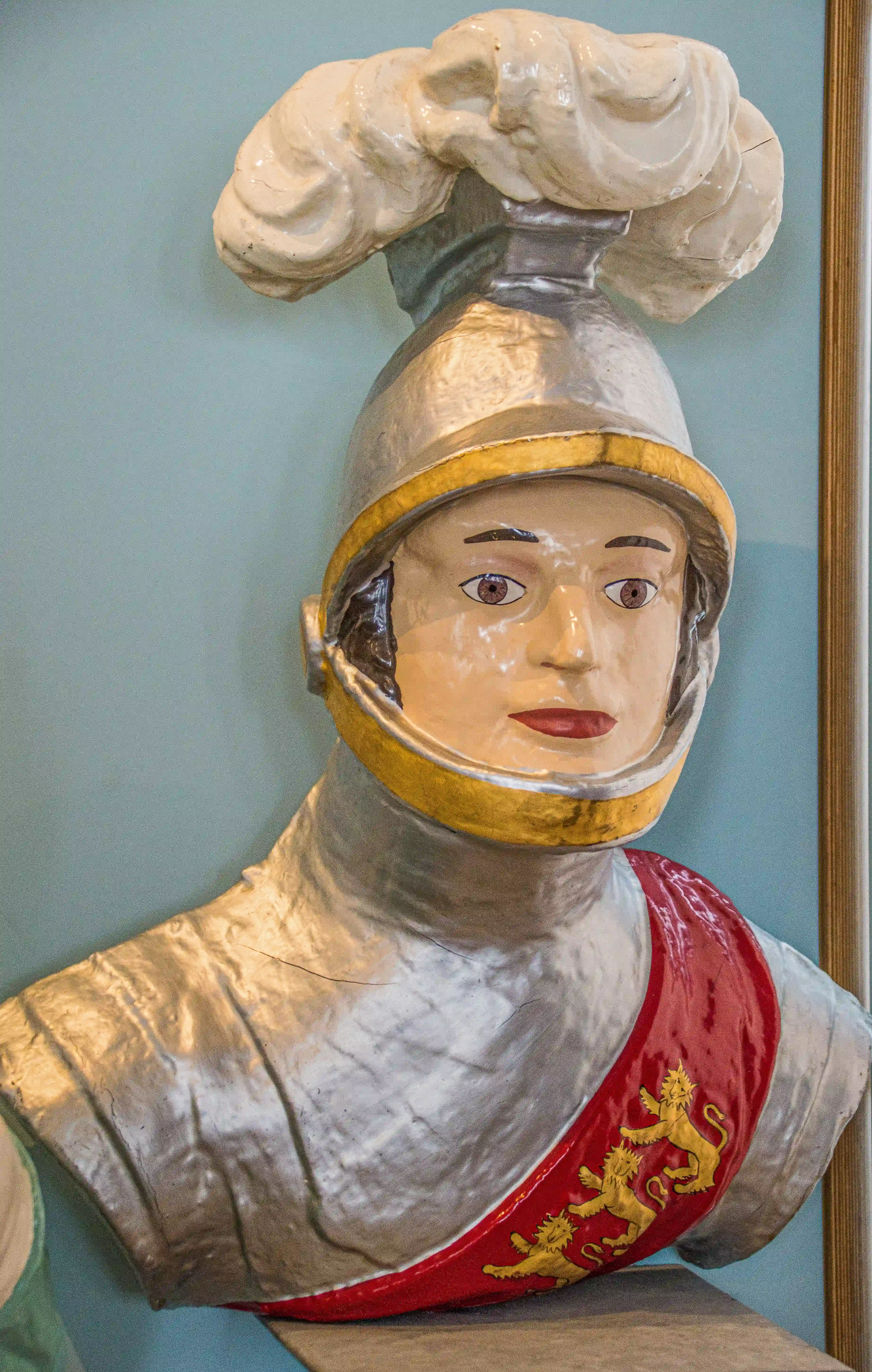 The image depicts a statue of a knight in armor, wearing a helmet with a yellow plume. The knight has a red sash with three golden lions, symbolizing a heraldic emblem. The statue appears to be made of a material resembling papier-mâché and is displayed against a light blue background