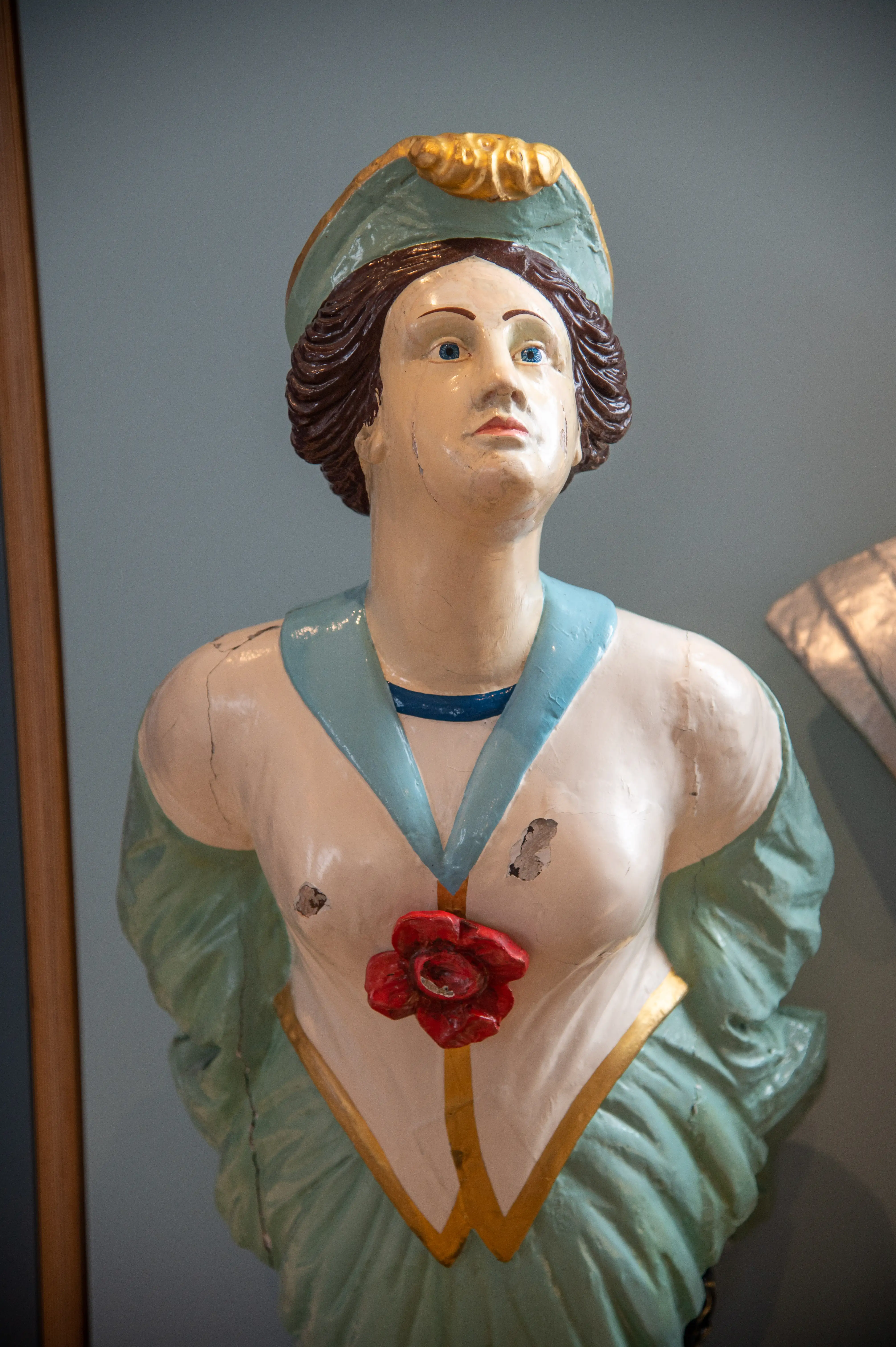 The image depicts a painted statue or figurine of a woman. The woman has a fair complexion, blue eyes, and brown hair, which is partially covered by a green headscarf with a yellow roll at the front. She is wearing a green dress with a low neckline, adorned with a red flower. The statue appears to be made of a material like papier-mâché or plaster and shows some signs of wear and damage.