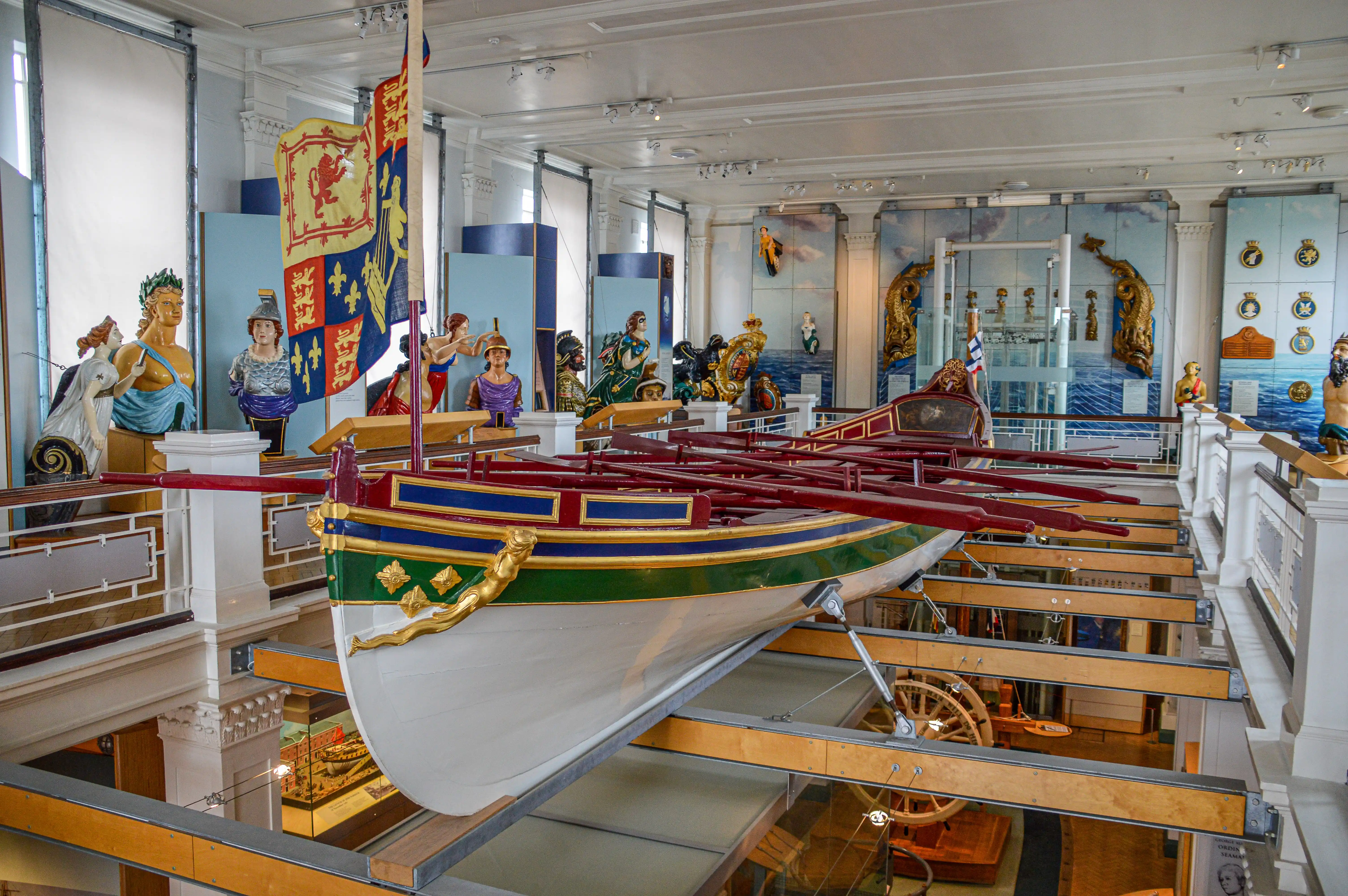 The image depicts a museum exhibit featuring a detailed model of an old sailing ship. The model is intricately designed with vibrant colors and gold accents, showcasing the ship's structure and decorations. Surrounding the ship are various maritime artifacts and statues, including figures in historical costumes and naval uniforms. The exhibit is well-lit, with spotlights highlighting different sections of the display. The background features blue and white wall panels, adding to the nautical theme of the exhibit.