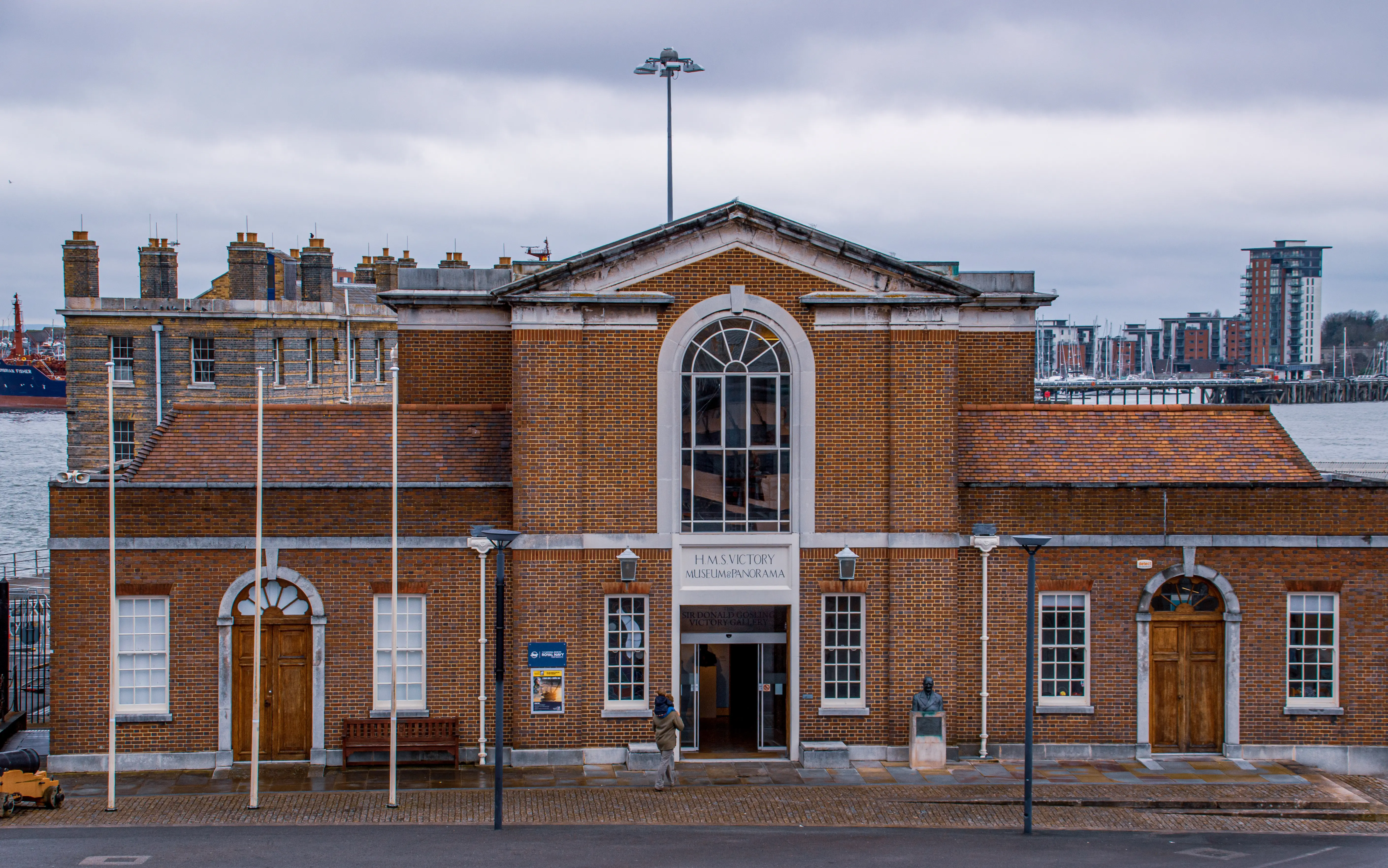 The image depicts a historic brick building located by the waterfront, identified as the HMS Victory Museum & Panorama. The structure features a prominent arched window and several wooden doors. A statue stands near the entrance, and the surrounding area includes modern buildings and a harbor with ships. The sky is overcast, suggesting a cloudy day.