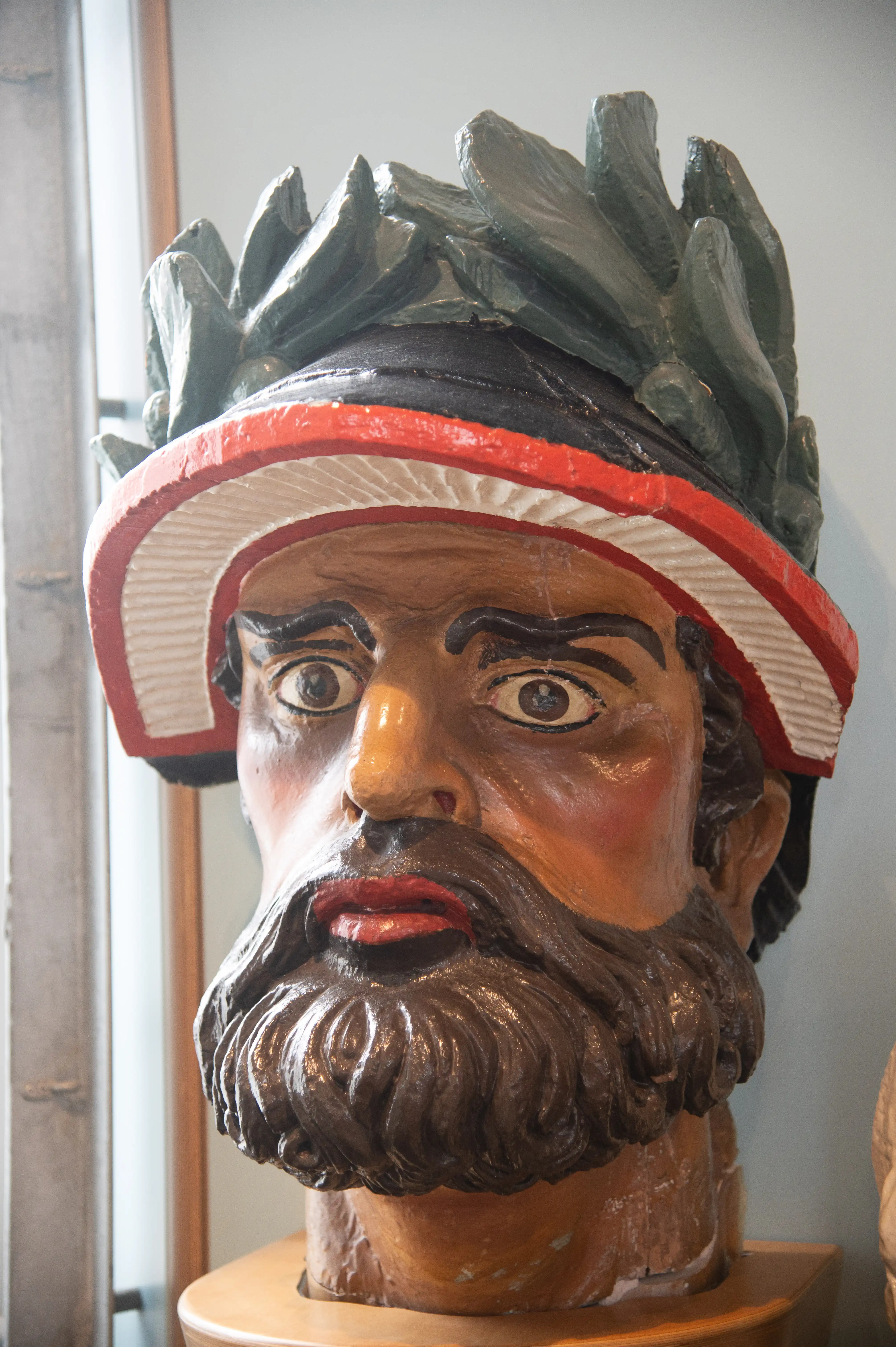 he image shows a detailed and colorful sculpture of a bearded man's head wearing a hat. The sculpture has expressive eyes, a prominent nose, and a red mouth. The hat is unique, featuring a combination of a straw hat and a green, leafy element on top.