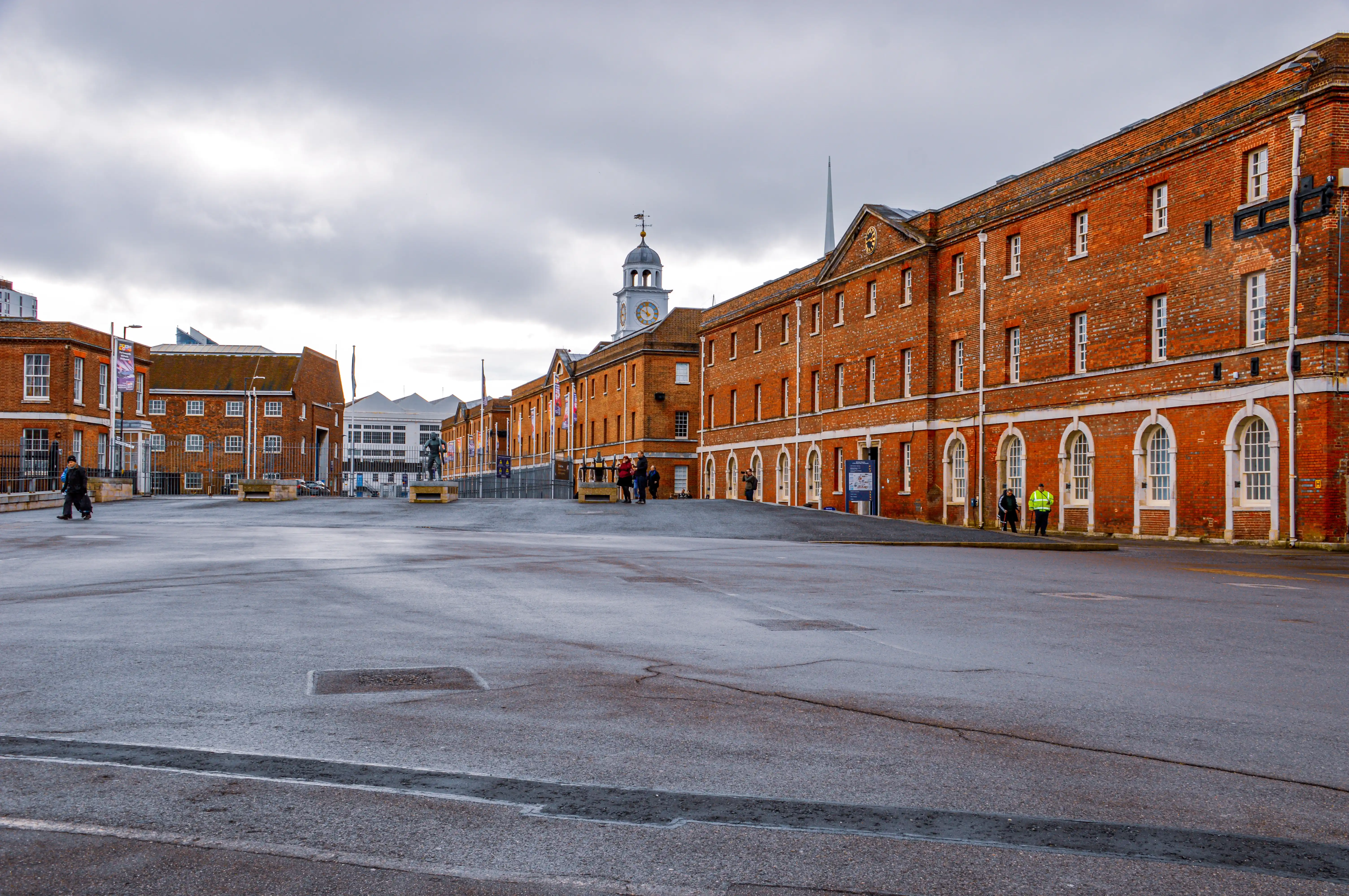 The image depicts a large, historic brick building with a clock tower, likely a former industrial or commercial structure now repurposed for modern use. The area is spacious and relatively empty, with a few people walking around. The sky is overcast, suggesting it might be a cold or rainy day. The architecture features large windows and arched doorways, giving it a classic and sturdy appearance. The overall scene is quiet and serene, with a sense of historical significance.
