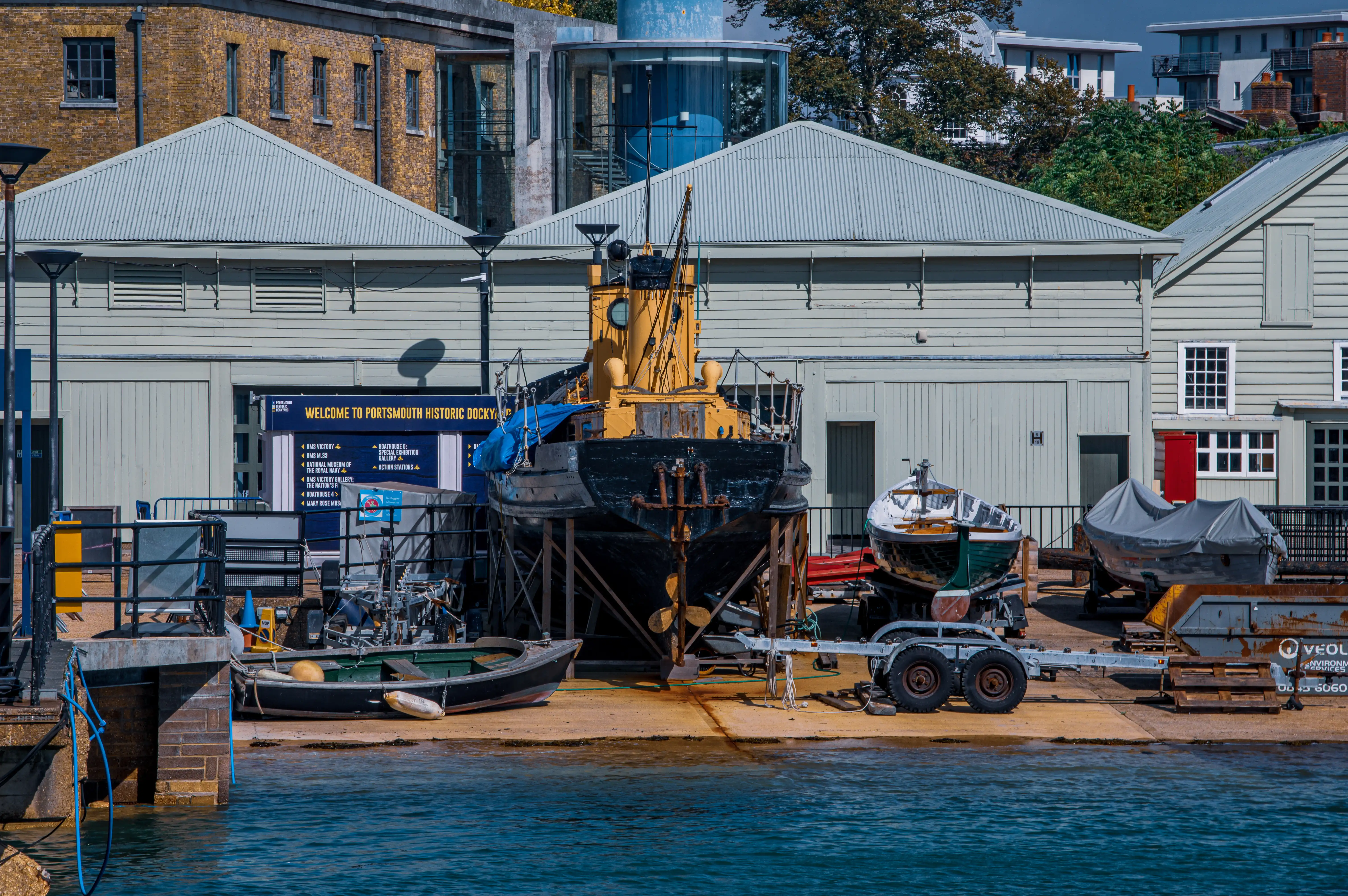 The image depicts Portsmouth Historic Dockyard, featuring various boats and a large ship under restoration or display. The dockyard buildings and other historic structures are visible in the background, along with a sign welcoming visitors to the site. The area appears to be a maritime museum or a historic site dedicated to preserving naval history.