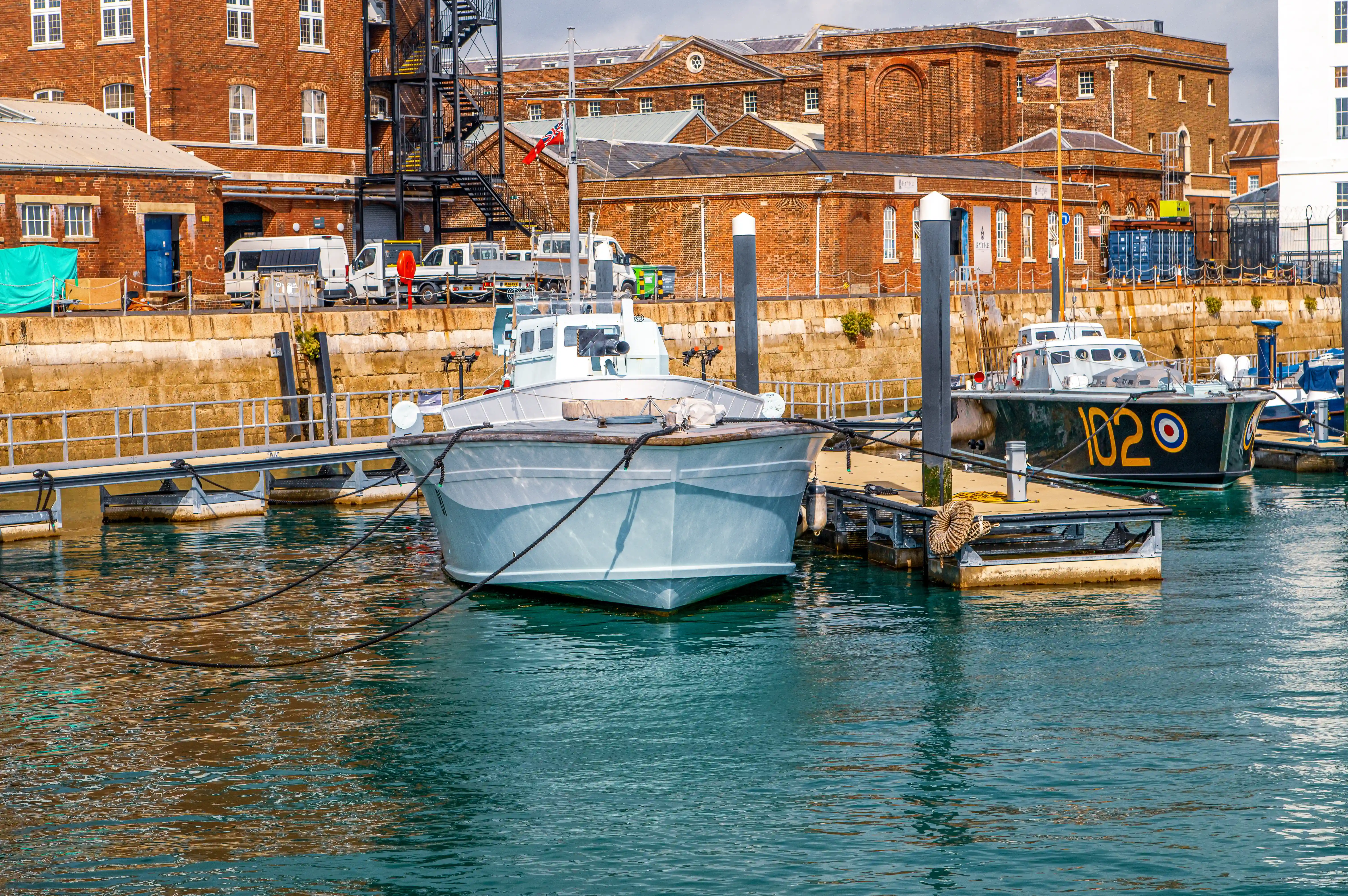 The image depicts a harbor scene with several boats docked at a pier. The boats are moored to concrete piers with ropes. In the background, there are historic brick buildings with large windows and some vehicles parked along the quay. The water in the harbor is calm, reflecting the boats and buildings. The overall atmosphere suggests a quiet, possibly commercial or industrial port area.