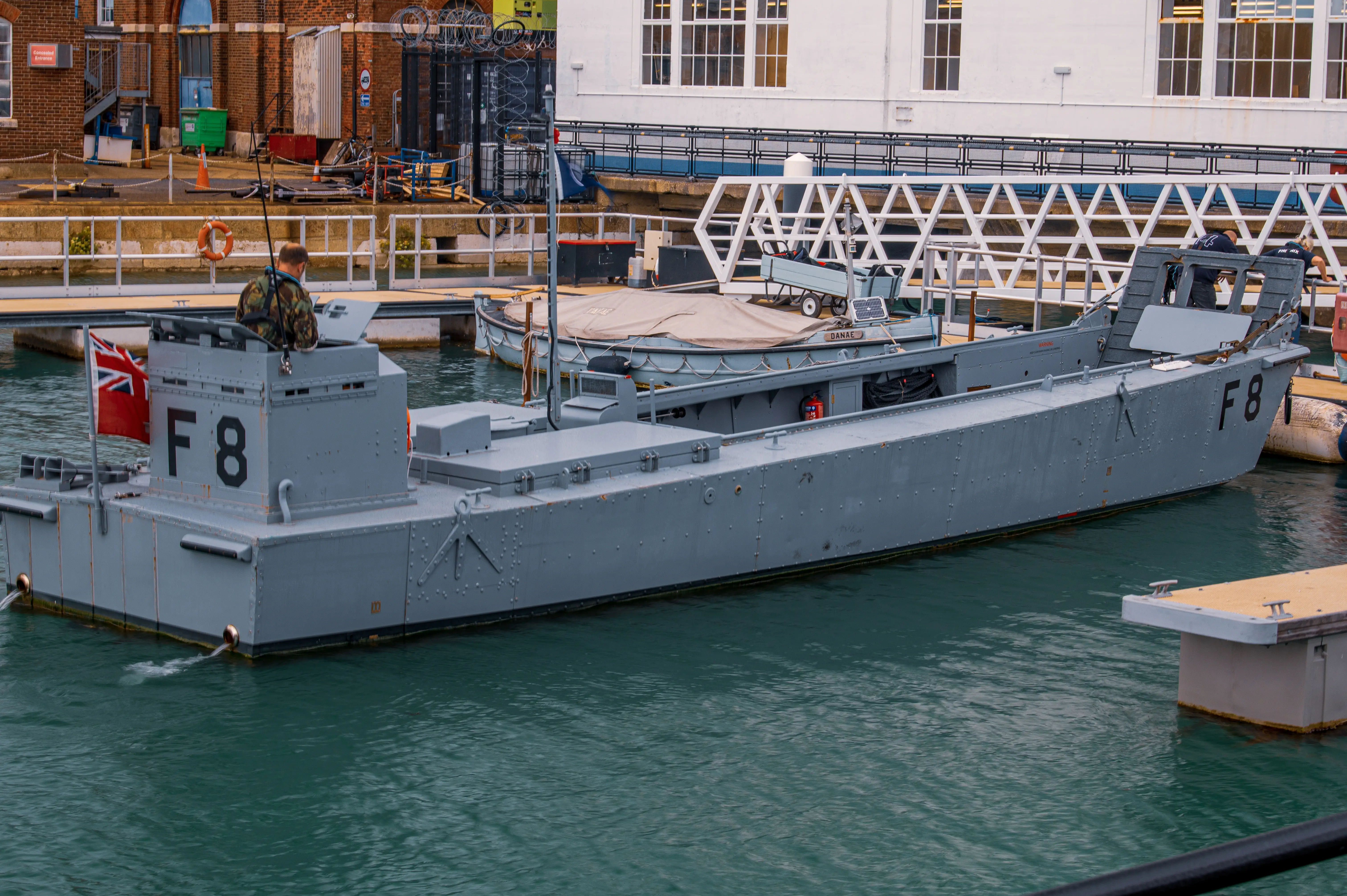 The image depicts a grey, military-style landing craft docked at a waterfront facility. The craft is marked with the identification 'F8' and features a person in camouflage clothing on board. The surrounding area includes other boats, a dock, and a building with large windows in the background.
