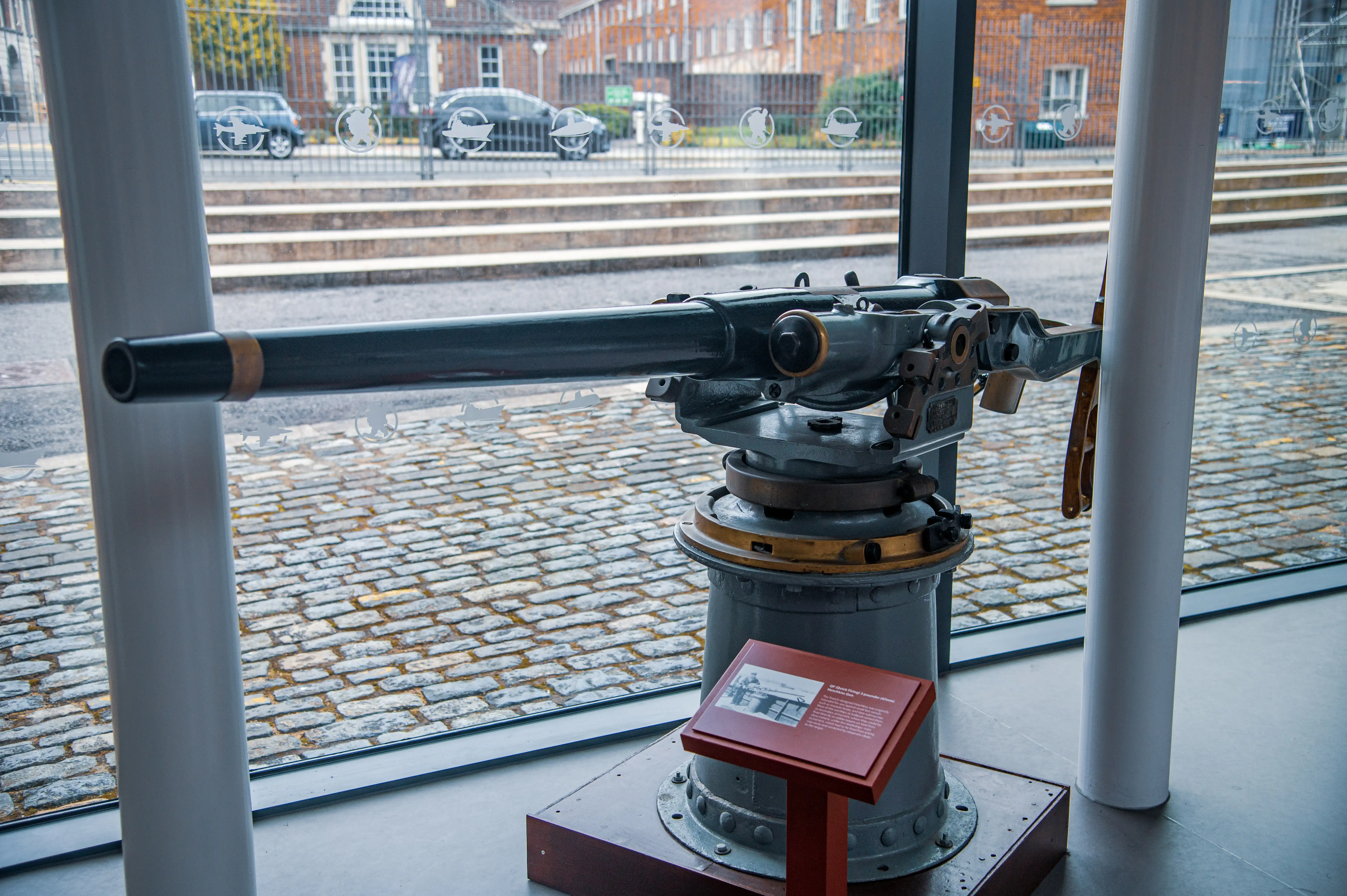The image shows a historical machine gun mounted on a tripod, displayed indoors near a window with a view of a cobblestone street and buildings outside. The machine gun is accompanied by an informational plaque, suggesting it is part of a museum exhibit.