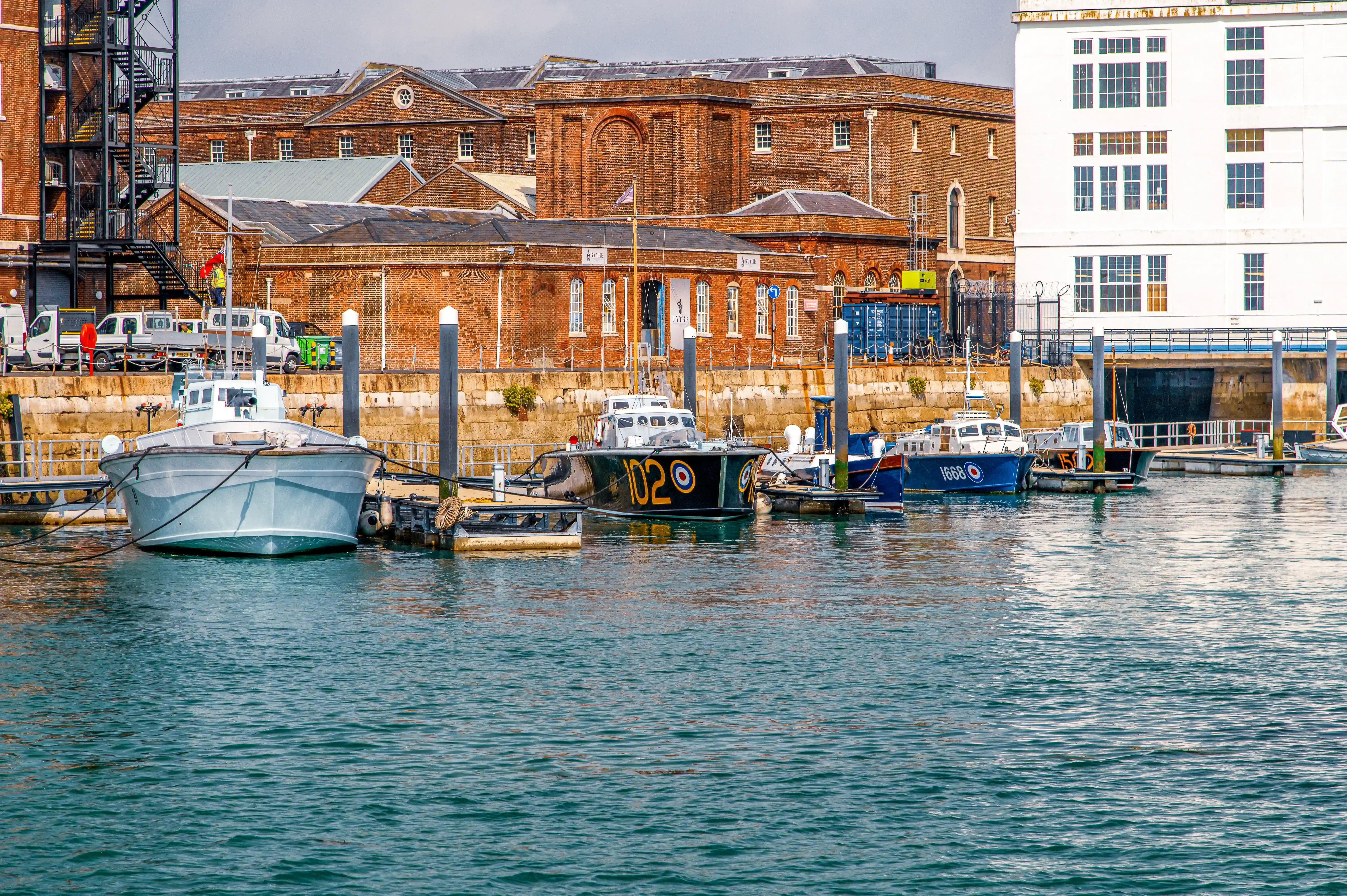 The image depicts a harbor scene with several boats docked at a pier. In the background, there are historic brick buildings with large windows and some modern elements like a fire escape on the left side. The water is calm, and the overall atmosphere suggests a quiet, possibly industrial or maritime area.