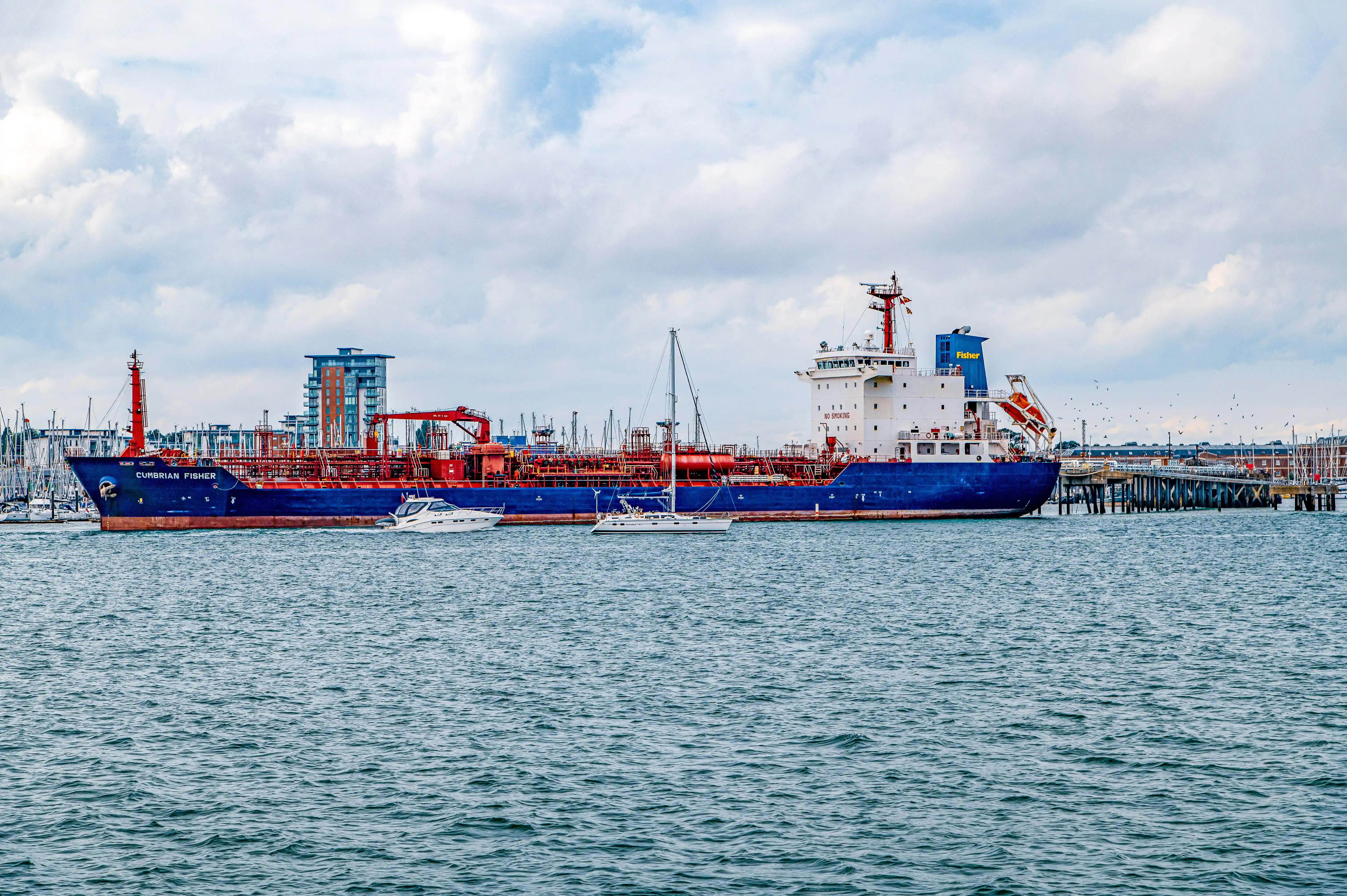 The image depicts a large cargo ship named 'Cumbrian Fisher' docked at a port. The ship is blue with red and white accents and is equipped with various cranes and industrial equipment. The port features modern buildings and infrastructure, including cranes and other industrial structures. Several smaller boats are seen near the ship, and the sky is partly cloudy. The overall scene suggests a busy, industrial maritime environment.