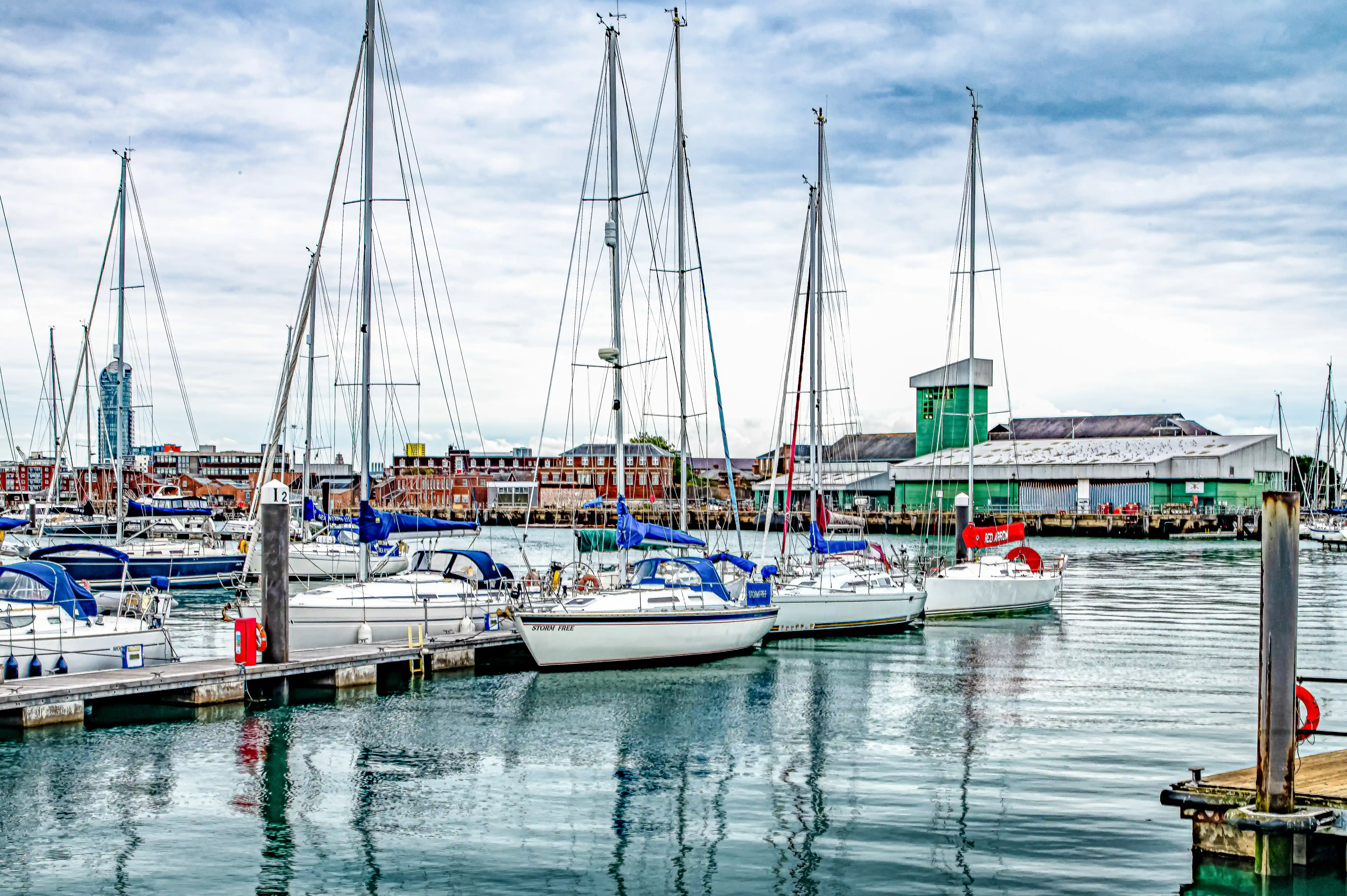 The image depicts a marina with several sailboats docked at the piers. The boats are moored in calm water, and their masts and rigging are clearly visible. In the background, there are various buildings, including a distinctive green structure and a tall tower. The sky is overcast, suggesting a cloudy day. The overall scene is serene and well-organized, typical of a marina setting.