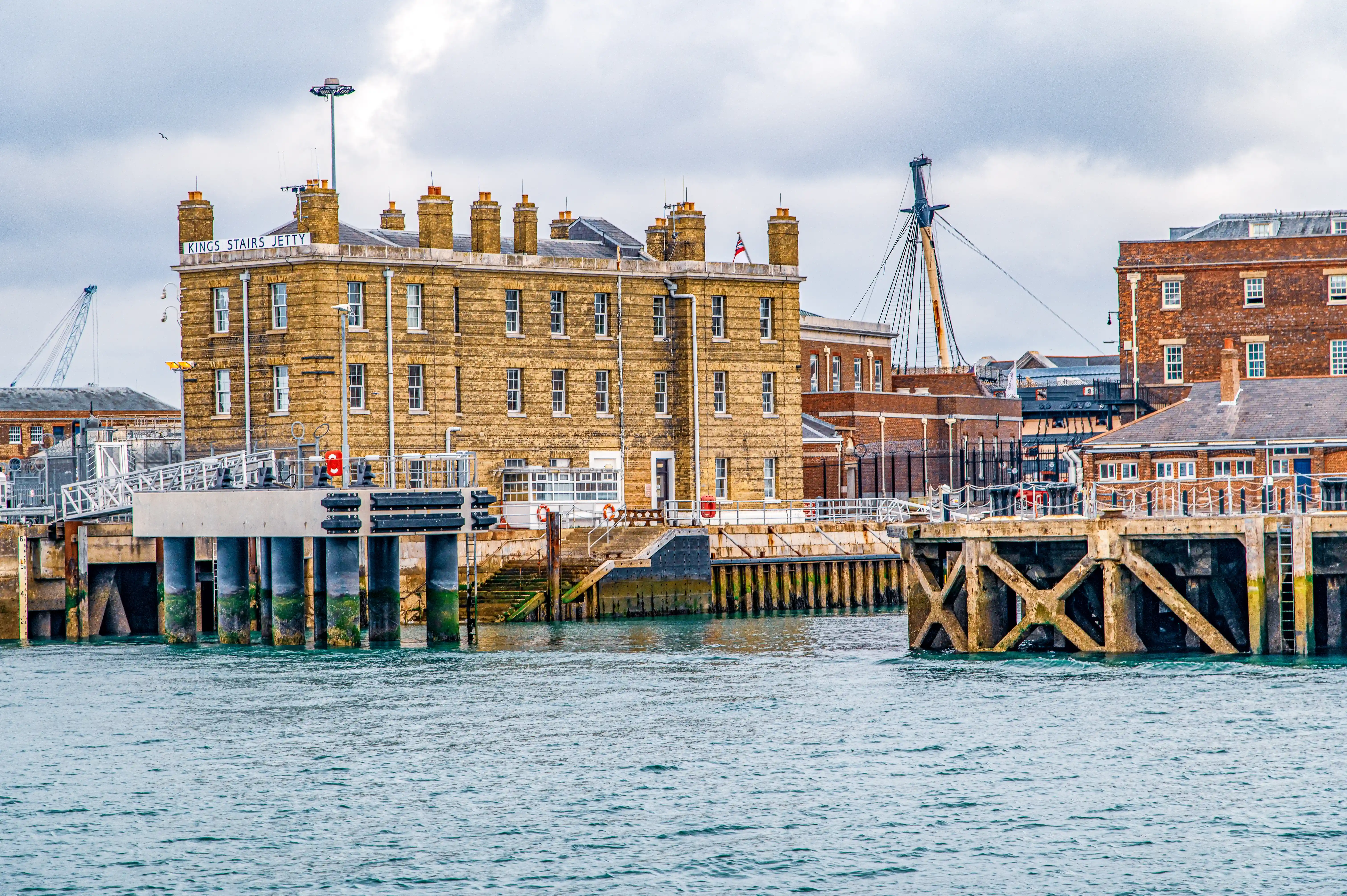 The image depicts a waterfront scene featuring a historic building labeled 'Kings Stairs Jetty.' The structure is made of stone with multiple chimneys and is situated next to a body of water. There is a wooden pier extending into the water, equipped with railings and life preservers. The sky is overcast, and there are various industrial elements in the background, including cranes and other buildings.