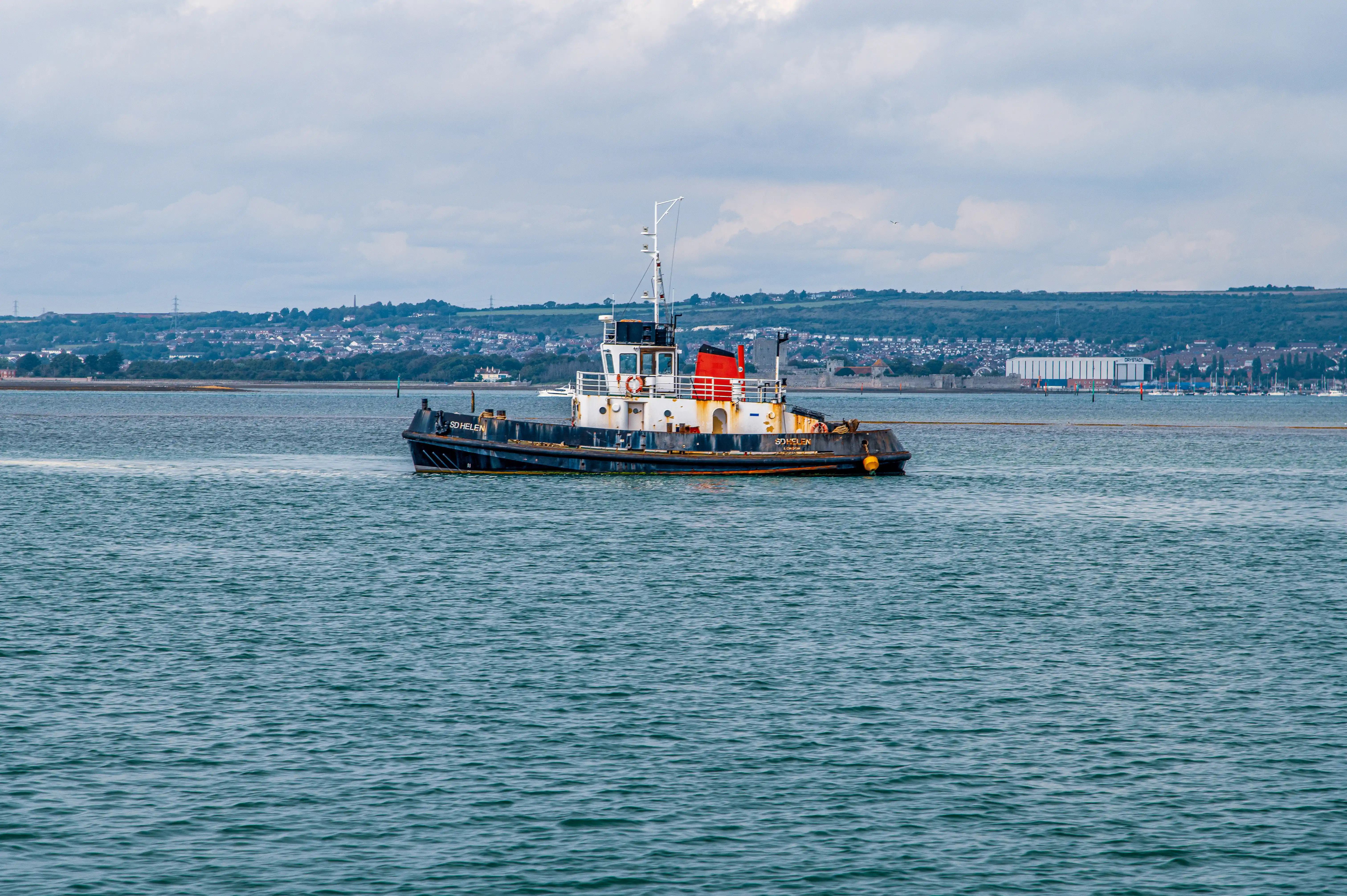 The image shows a tugboat named 'SD HELEN' sailing in a body of water. The boat is painted in dark blue with a white superstructure and has a red and white mast. The background features a shoreline with various buildings, greenery, and a partly cloudy sky.