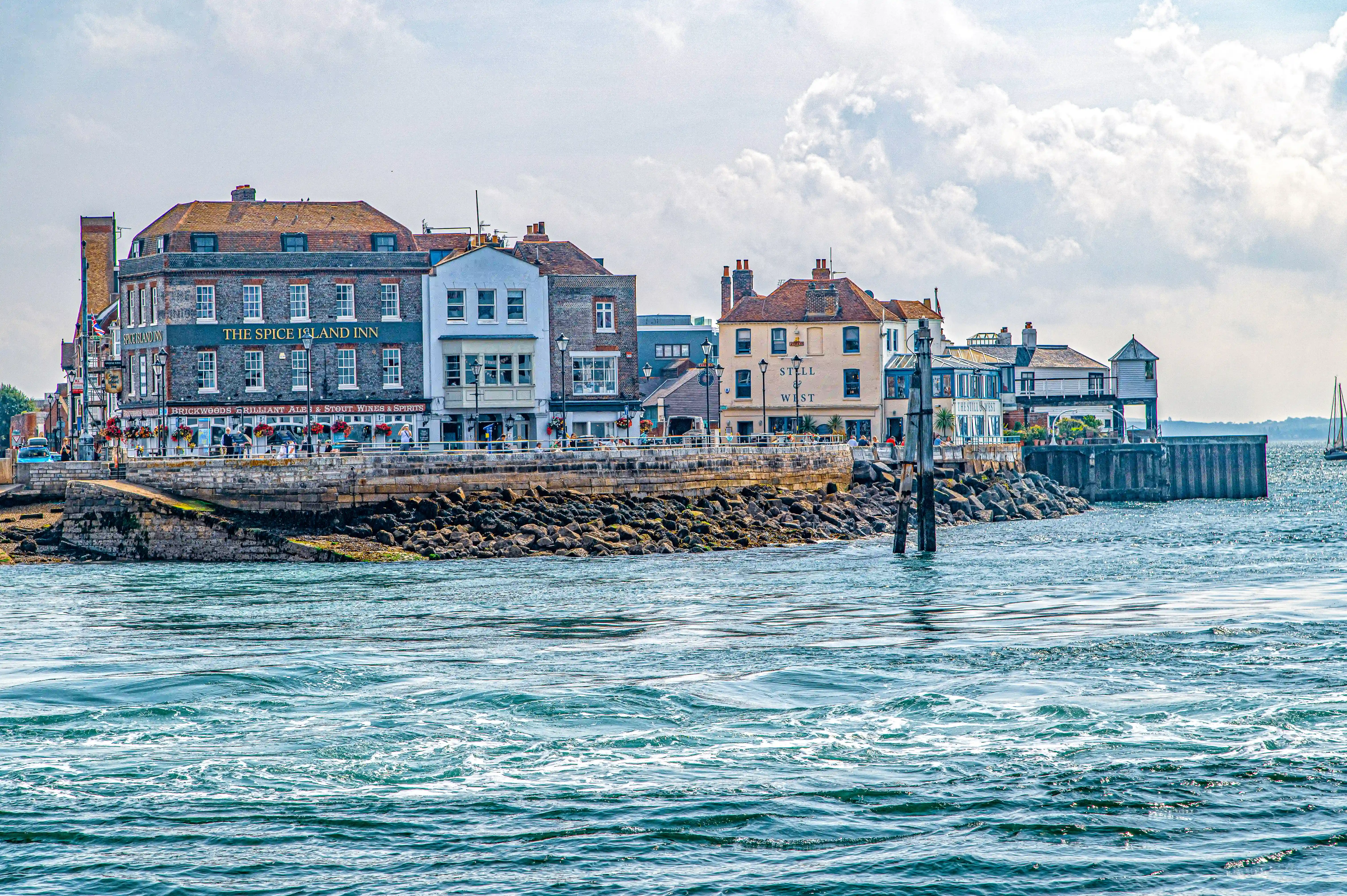 The image depicts a picturesque waterfront scene featuring a row of charming, historic buildings. Prominent establishments include 'The Spice Island Inn' and 'Still & West.' The buildings are situated along a stone-paved promenade, adorned with flower baskets and street lamps. The waterfront is lined with a stone breakwater, and the scene is set against a backdrop of a cloudy sky and calm water.