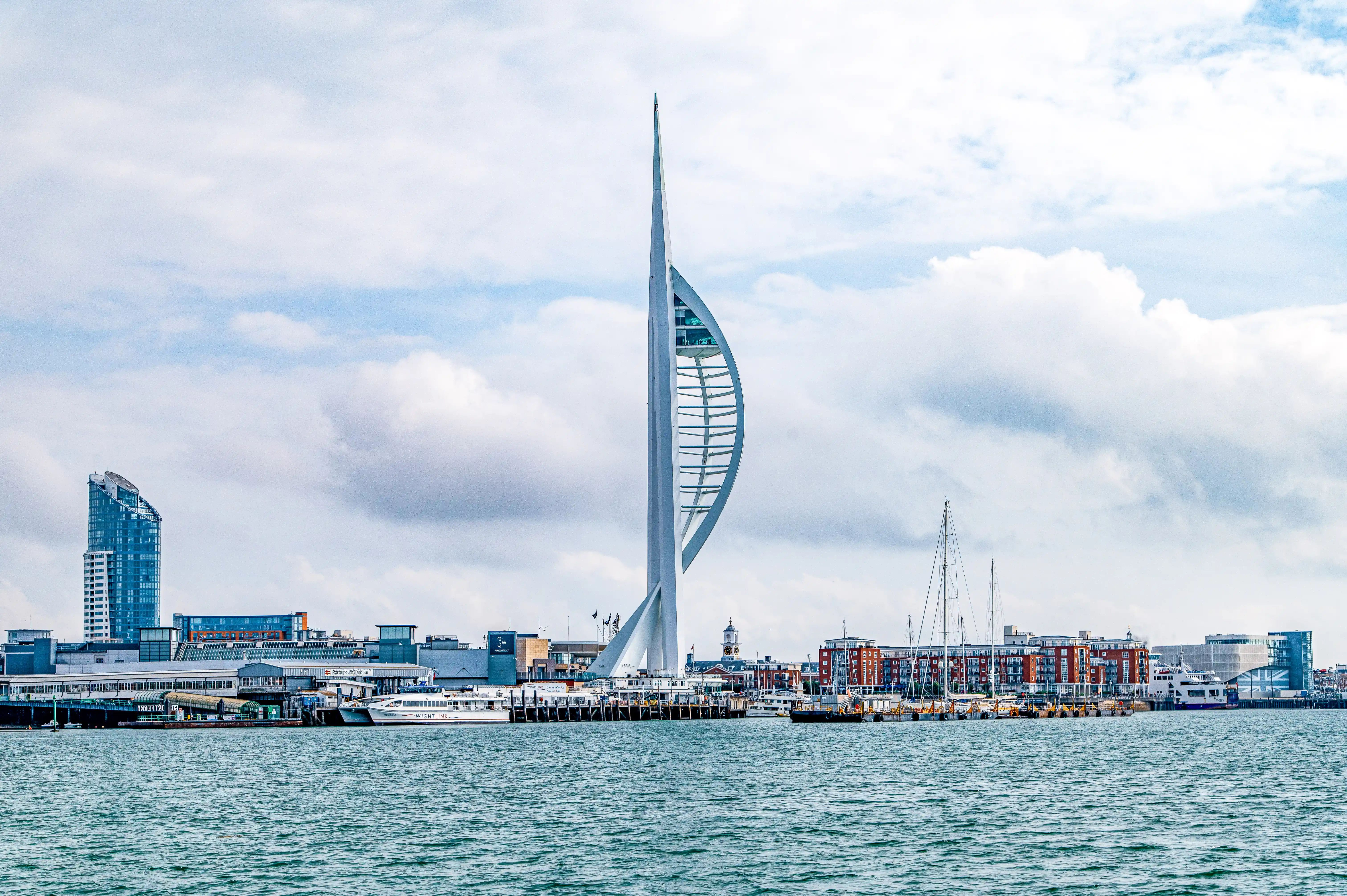 The image depicts a waterfront cityscape featuring a prominent tall, modern structure known as the Spinnaker Tower, which resembles a sail. The tower is situated near a harbor with several boats and yachts docked. The surrounding area includes a mix of modern buildings and older, brick structures, indicating a blend of contemporary and historical architecture. The sky is partly cloudy, and the water in the harbor is calm.