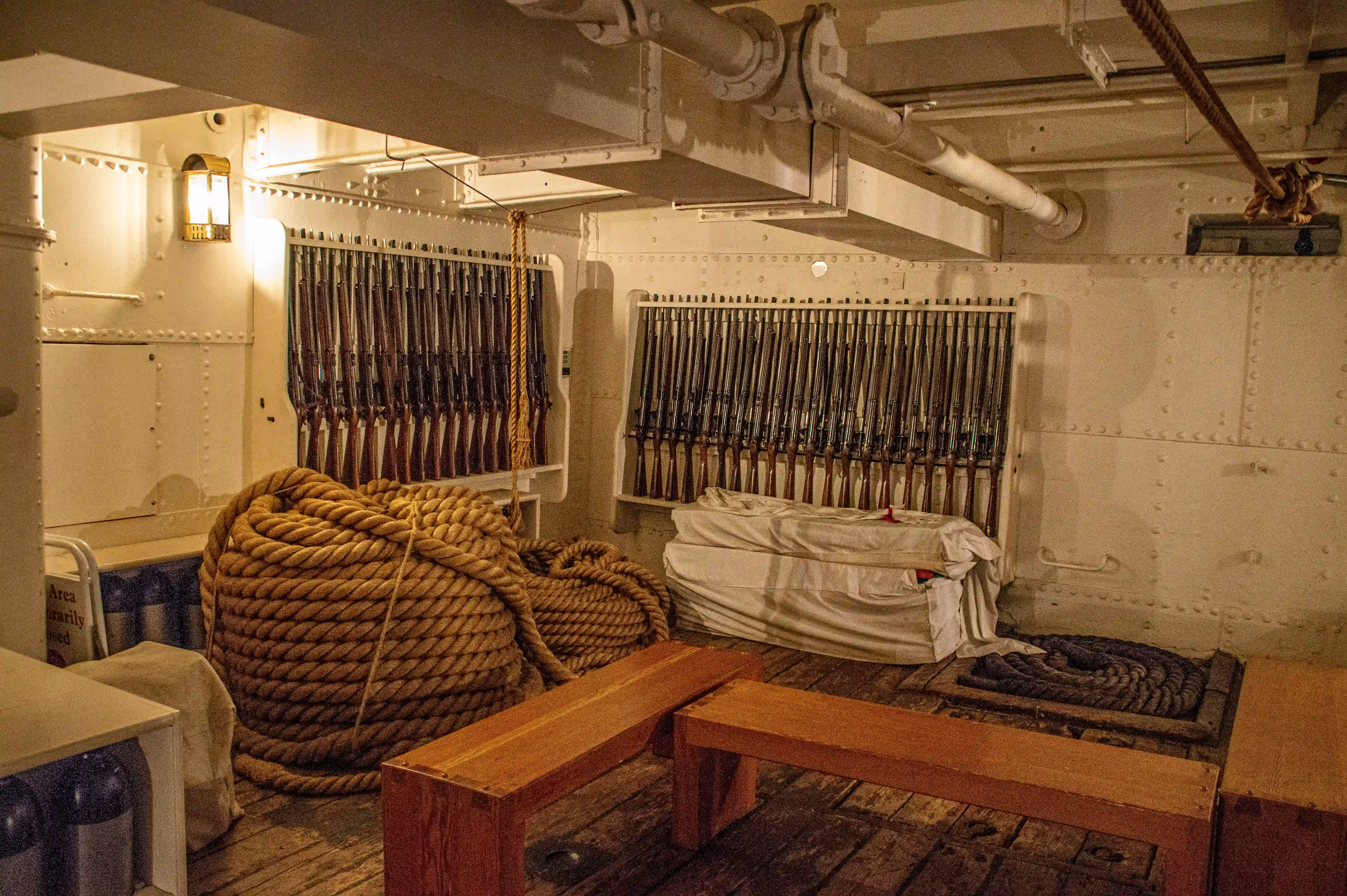 The image depicts the interior of a ship, specifically an area that appears to be a storage or utility room. The room contains various maritime tools and equipment, including multiple boats hooks hanging on racks, coils of rope, covered objects that might be additional tools or supplies, and wooden boxes. The setting suggests a well-organized space designed for easy access to necessary items during maritime activities.