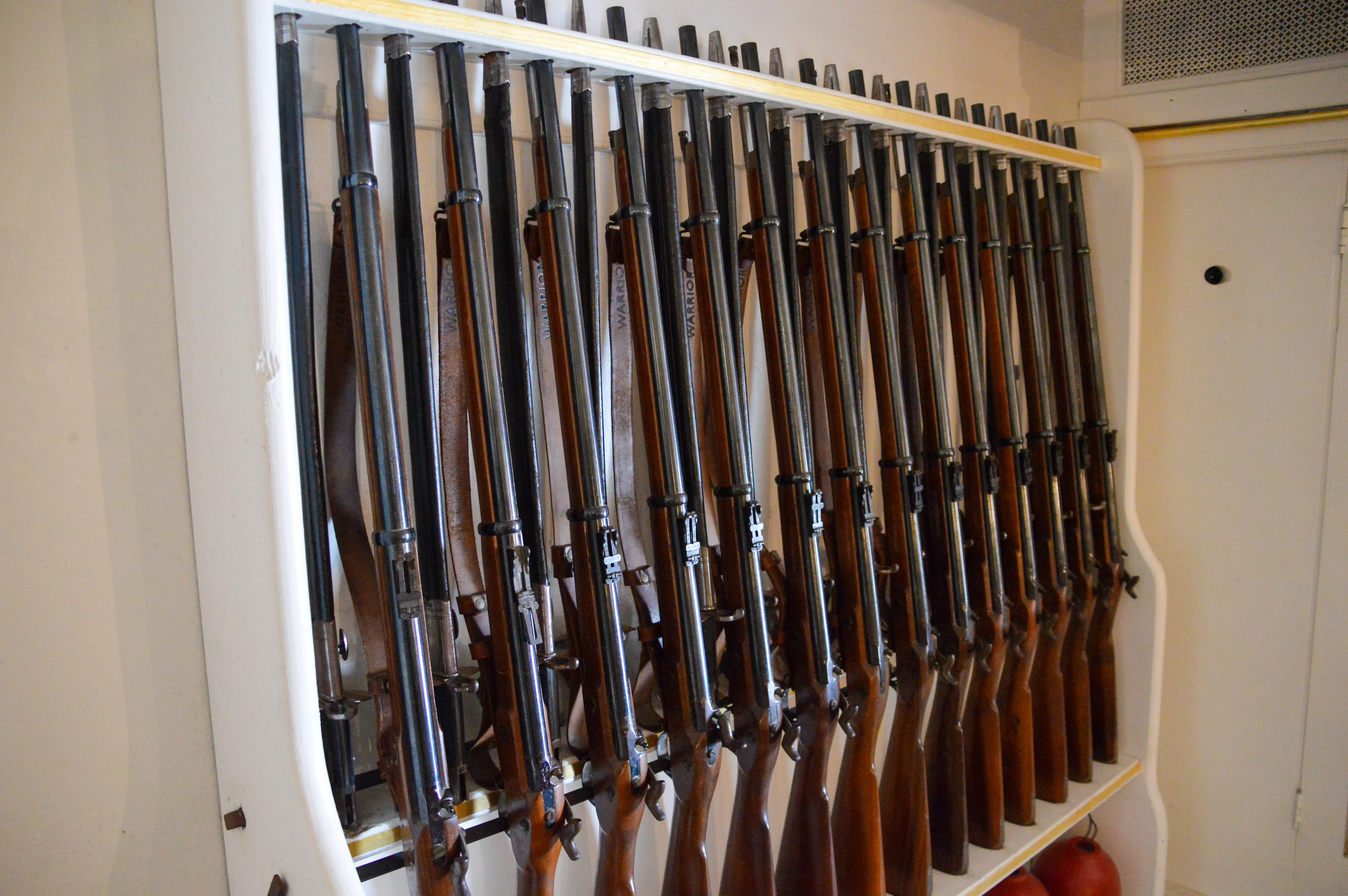 The image shows a collection of rifles neatly arranged in a vertical gun rack. The rifles have wooden stocks and appear to be of similar make and model, with the word 'Warrior' visible on some of the barrels. The rack is mounted on a wall and is designed to hold the rifles securely in place.