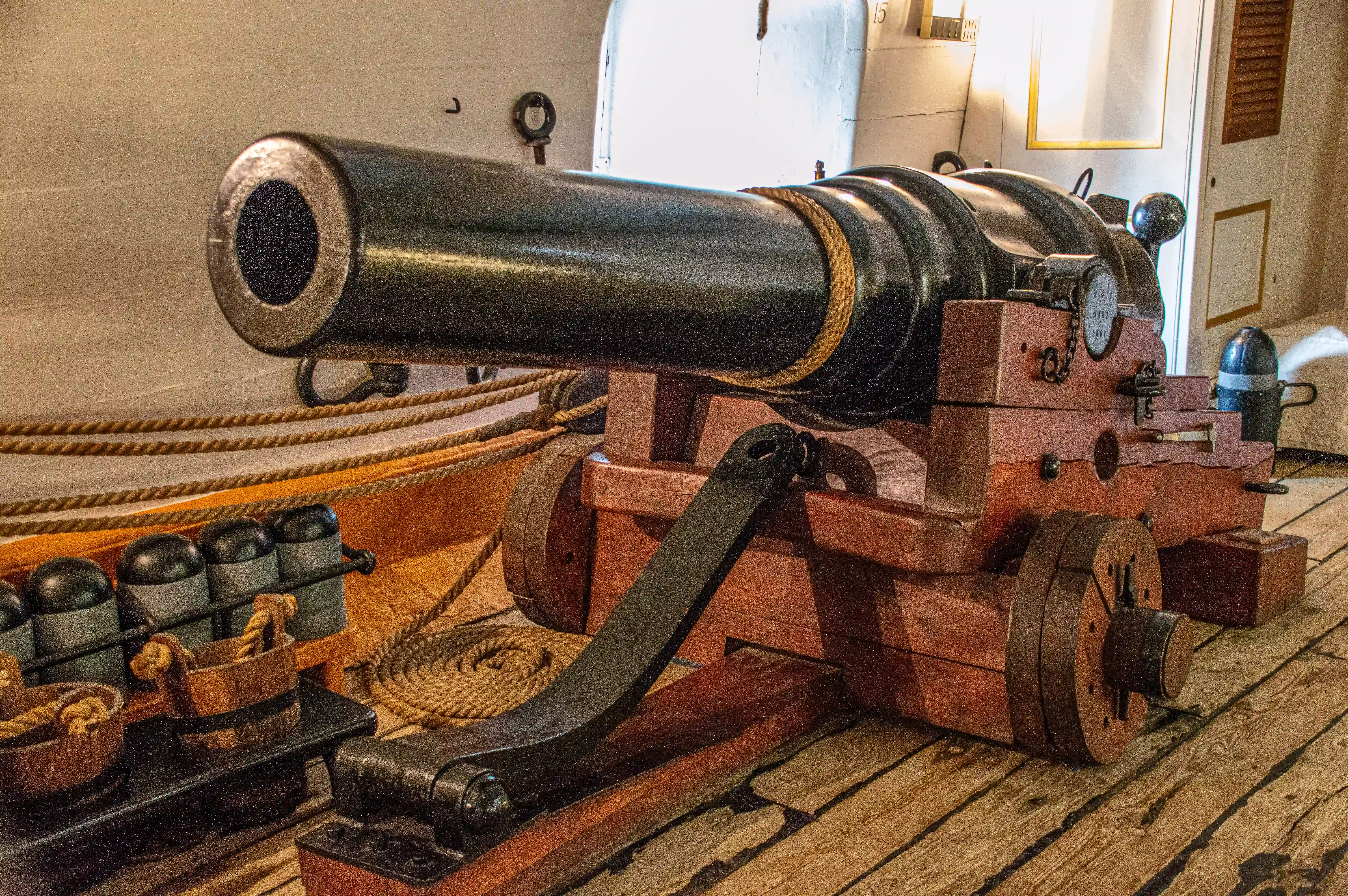 The image depicts an old naval cannon mounted on a wooden carriage, likely from a historical ship. The cannon is situated on a wooden deck, with various associated equipment such as cannonballs, wooden wedges, and ropes visible around it. The setting appears to be part of a museum or historical exhibit, showcasing the artillery used in naval warfare.