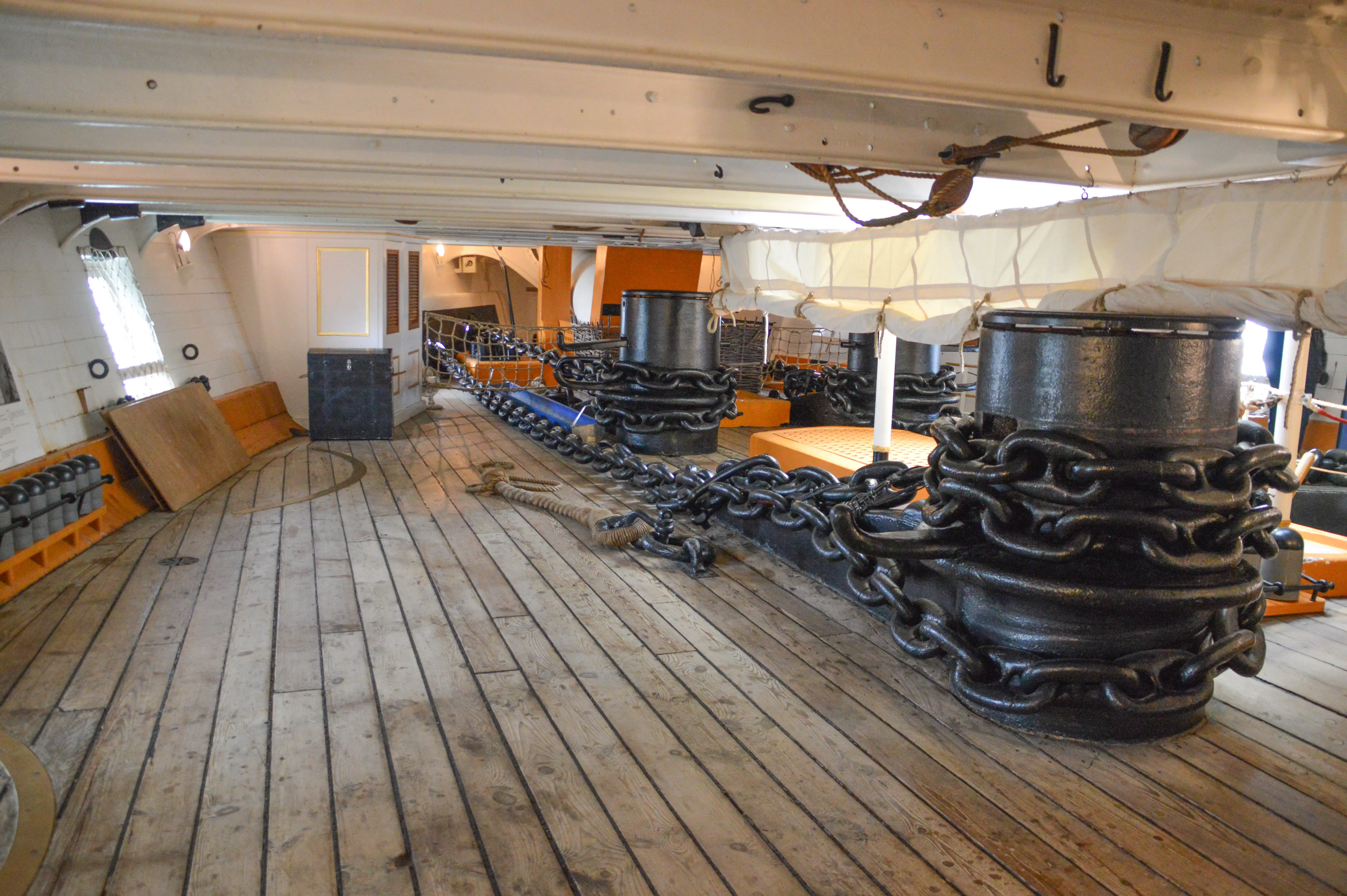 The image depicts the interior of an old ship, likely a historical warship, showing large black cannons secured with thick ropes and chains on a wooden deck. The ship's interior features wooden planks, white walls with portholes, and various naval equipment, giving a glimpse into maritime history and naval warfare.