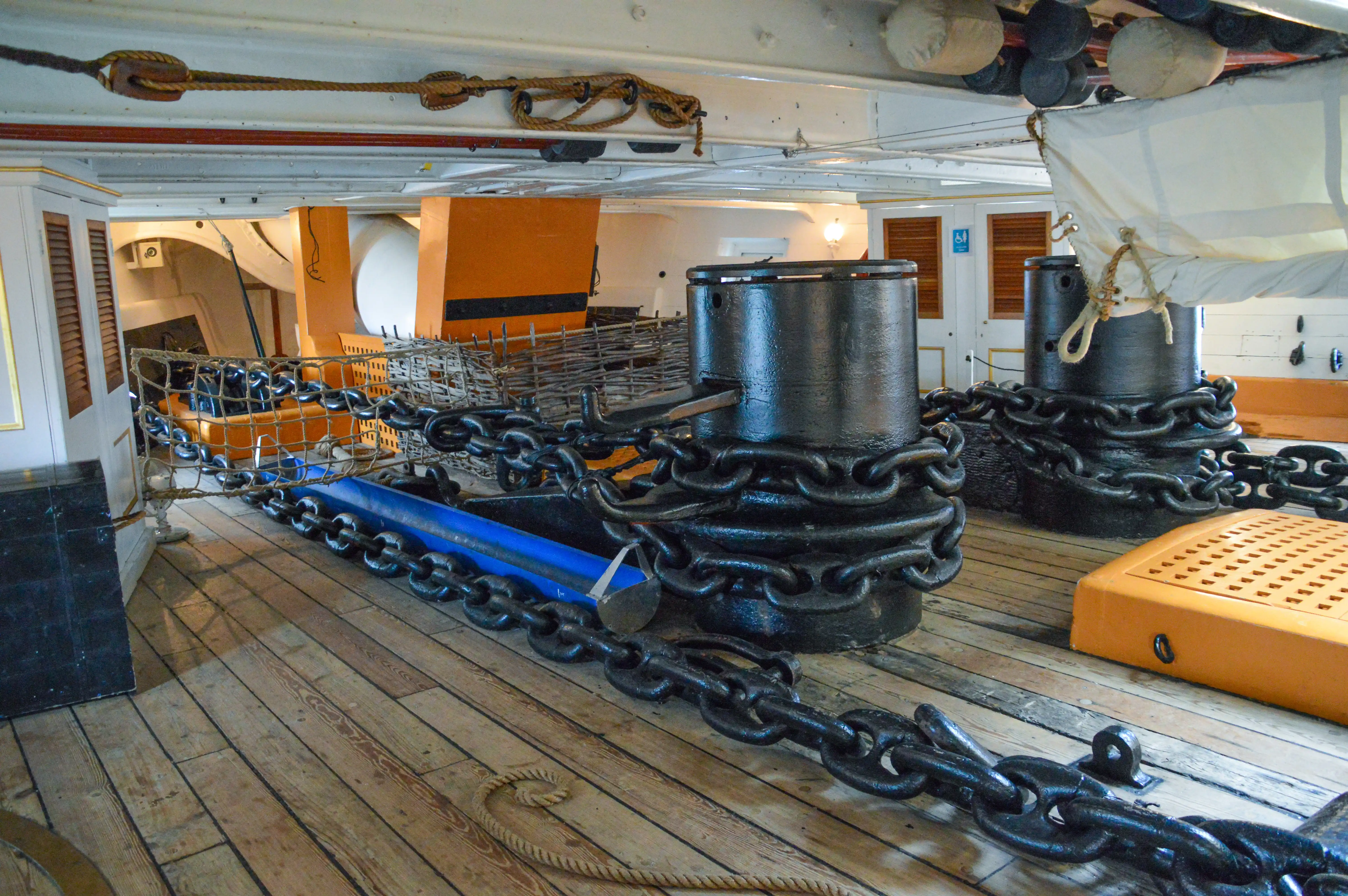 The image depicts the interior of a historical ship, showcasing large black cylindrical objects, likely cannons, secured with thick chains. The deck is made of wooden planks, and various ropes and rigging are visible overhead. The background includes orange-painted walls and wooden structures, suggesting the ship's interior compartments and storage areas.
