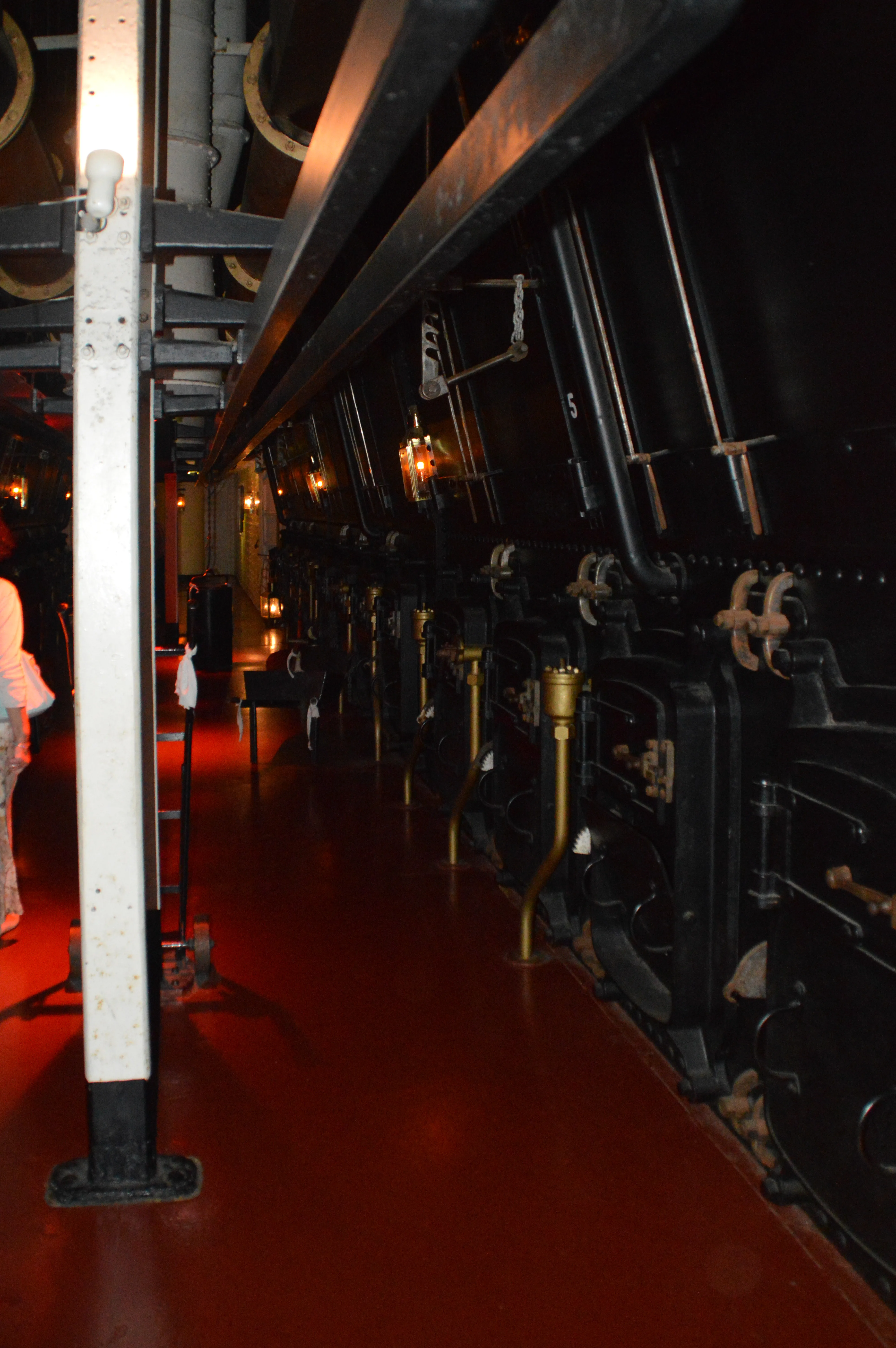 The image depicts the interior of a historical ship engine room, showcasing large, vintage machinery and equipment. The room is dimly lit with warm lighting, highlighting the intricate details of the engine components. The floor is red, and there are visible signs of wear and tear on the machinery, indicating its age. A person in a pink outfit is partially visible on the left side of the image, adding a sense of scale to the scene. The overall atmosphere is one of historical significance and industrial heritage
