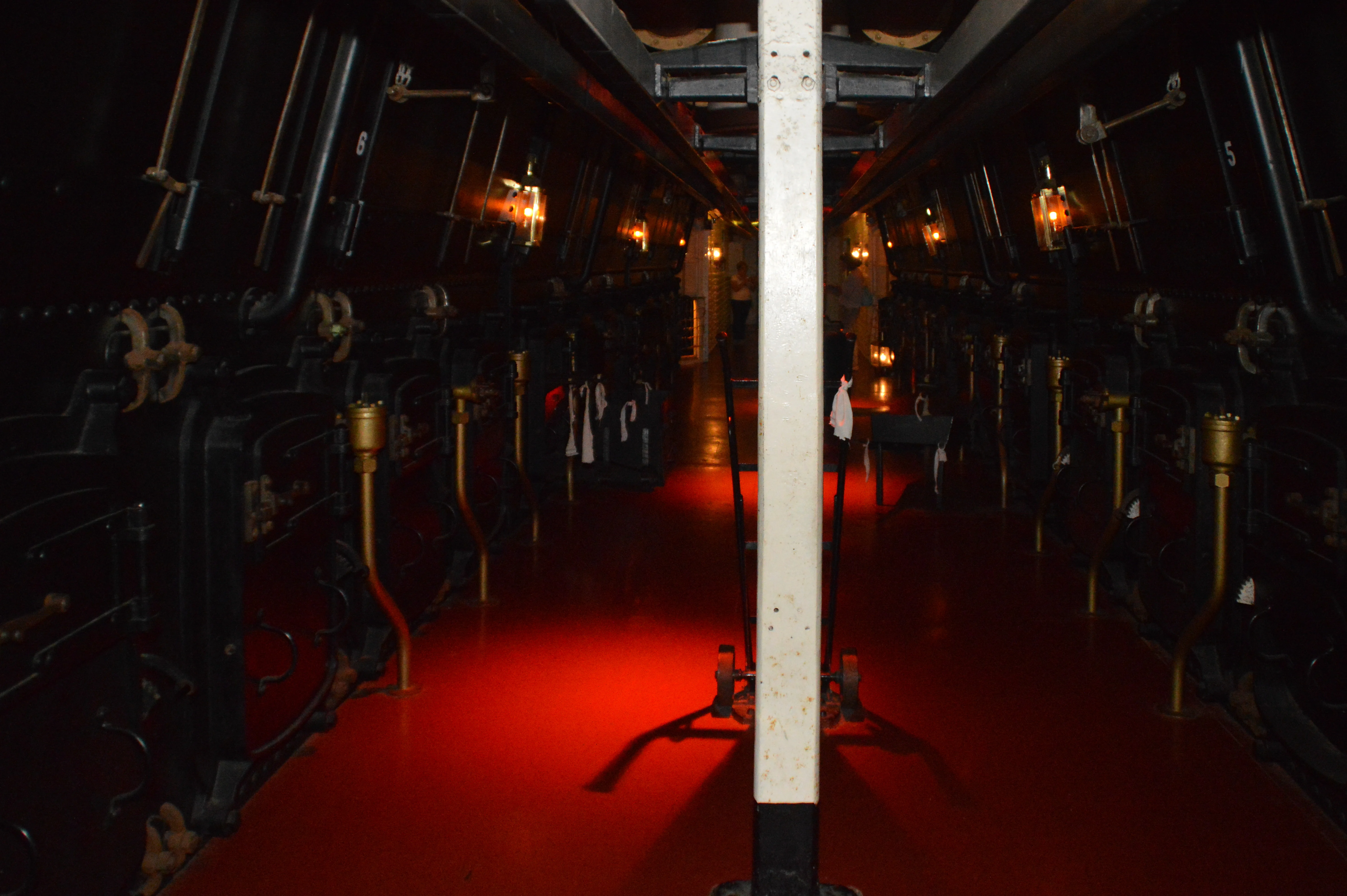 The image depicts an interior space with a red carpet and dim lighting. The room features multiple chairs with brass accents and black upholstery, arranged in rows on either side of a central aisle. The lighting is provided by wall-mounted lamps with a vintage design, casting a warm glow. The overall atmosphere appears intimate and somewhat formal, possibly suggesting a setting like a theater, lounge, or dining area.