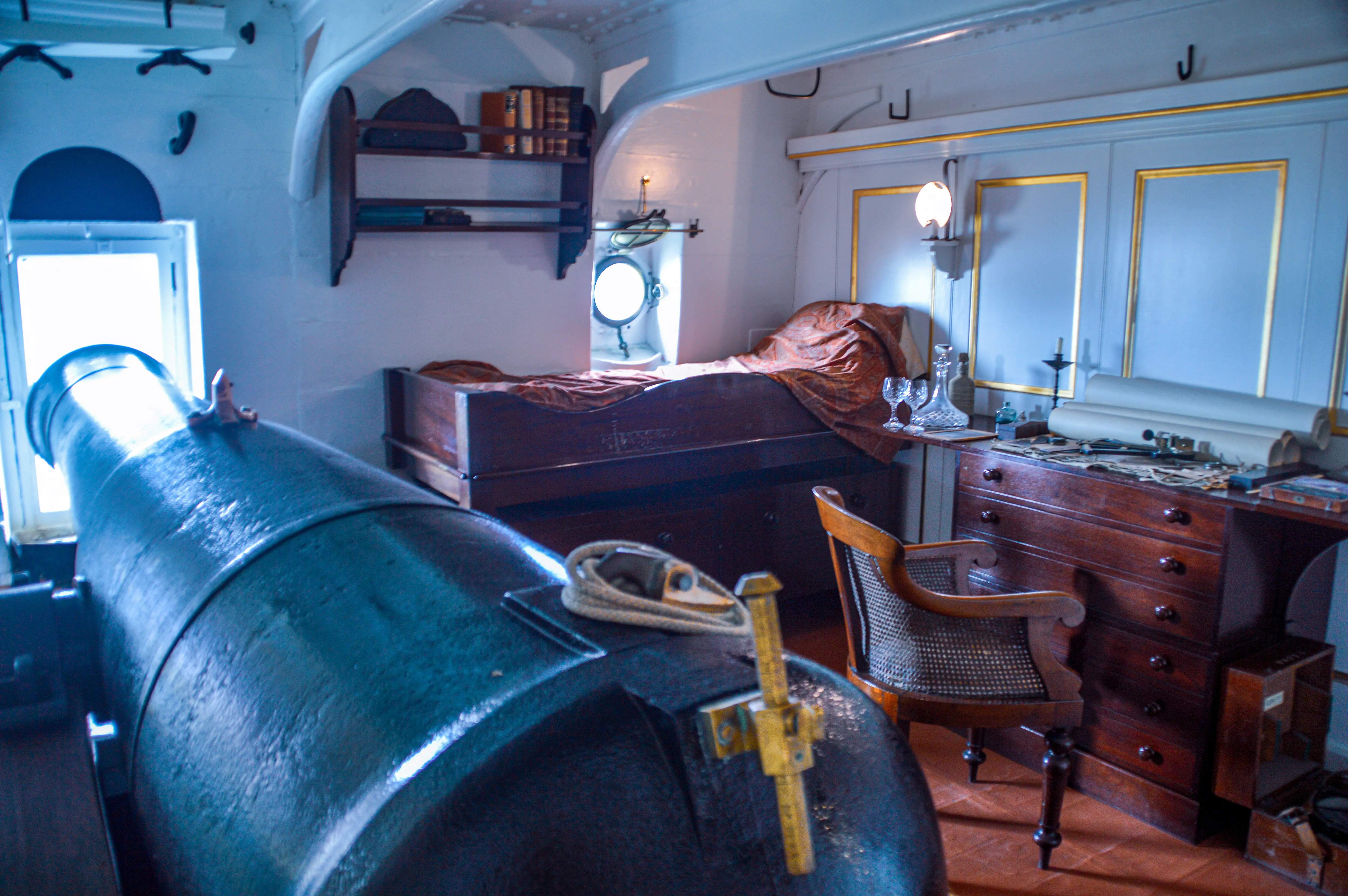 The image depicts a historical ship cabin interior with a neatly made bed, a wooden desk with various items, a wicker chair, and a porthole window allowing natural light to enter. The cabin has a cozy and organized appearance, with books and other personal items neatly arranged on shelves and the desk.