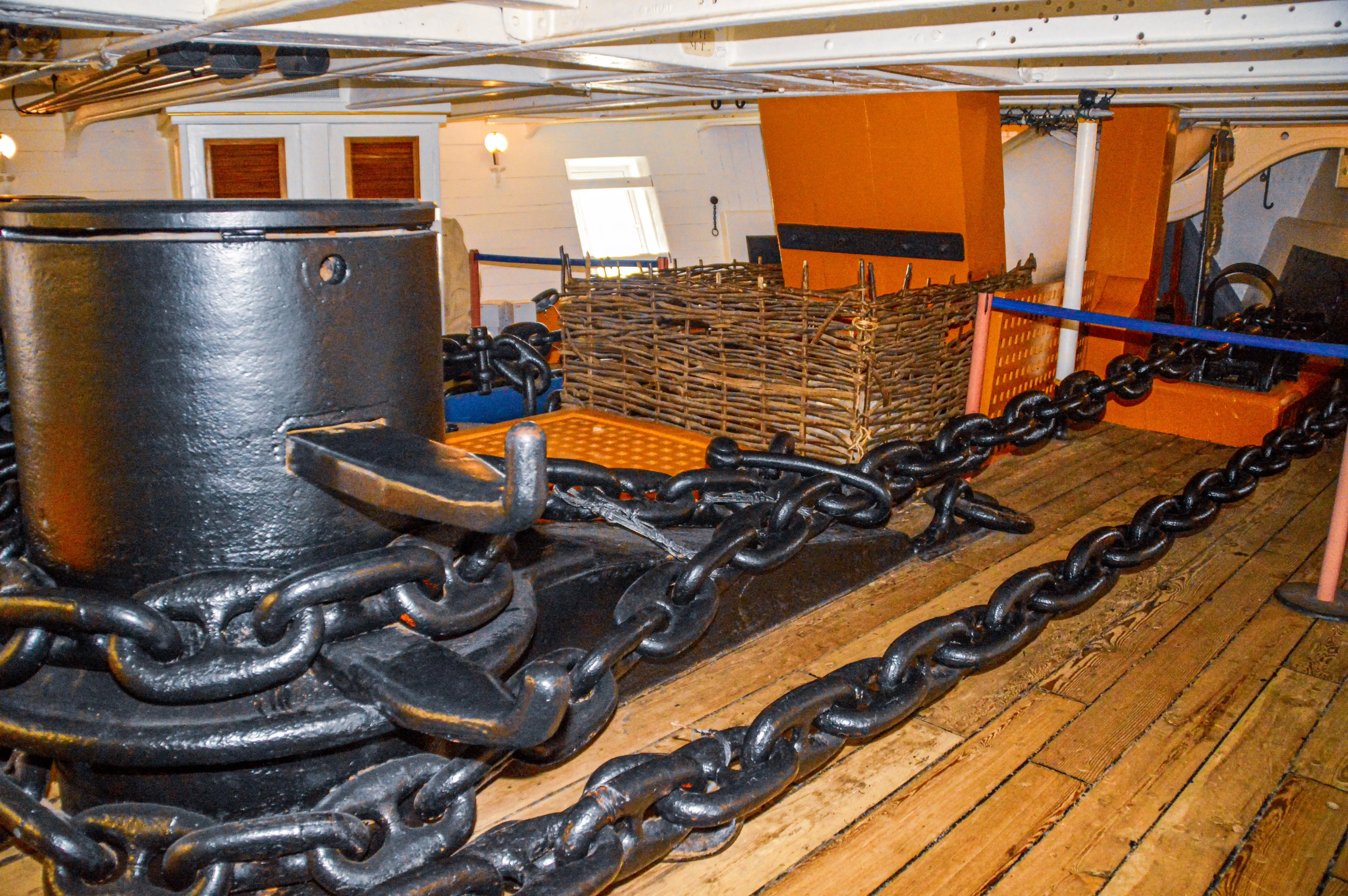 The image depicts the interior of an old ship, likely a warship, featuring large cannons secured with thick chains. The deck is made of wooden planks, and there are wicker barriers and orange structural elements visible in the background. The setting appears to be part of a museum exhibit, showcasing historical naval artifacts.