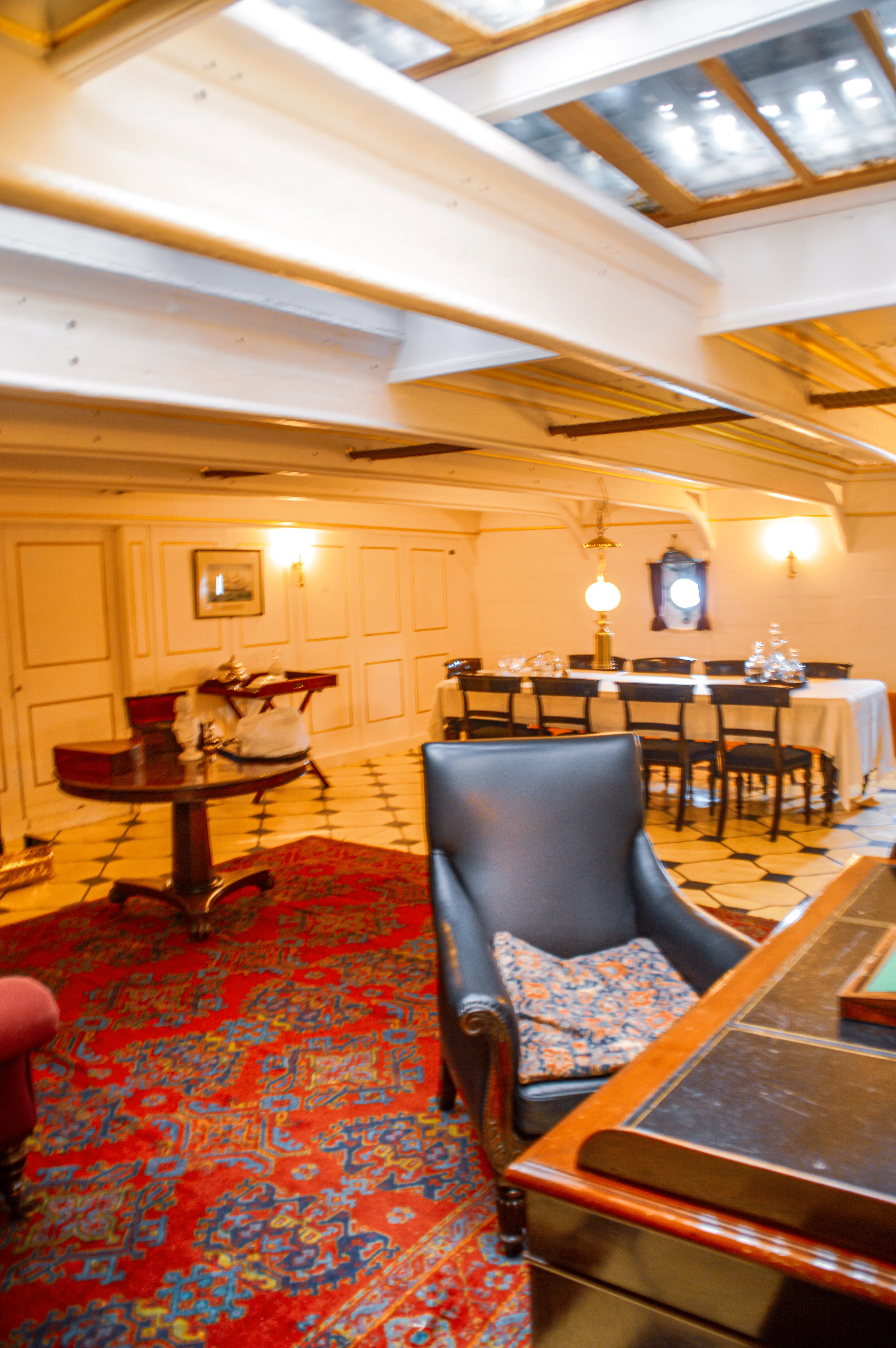 The image depicts an elegantly furnished room with wooden paneling, a patterned rug, and a mix of vintage furniture. The room features a cozy seating area with a blue leather chair and a small table with a lamp. In the background, there is a dining area set with tables and chairs, and a chandelier hanging from the ceiling. The overall ambiance is warm and inviting, suggesting a classic or historical setting.