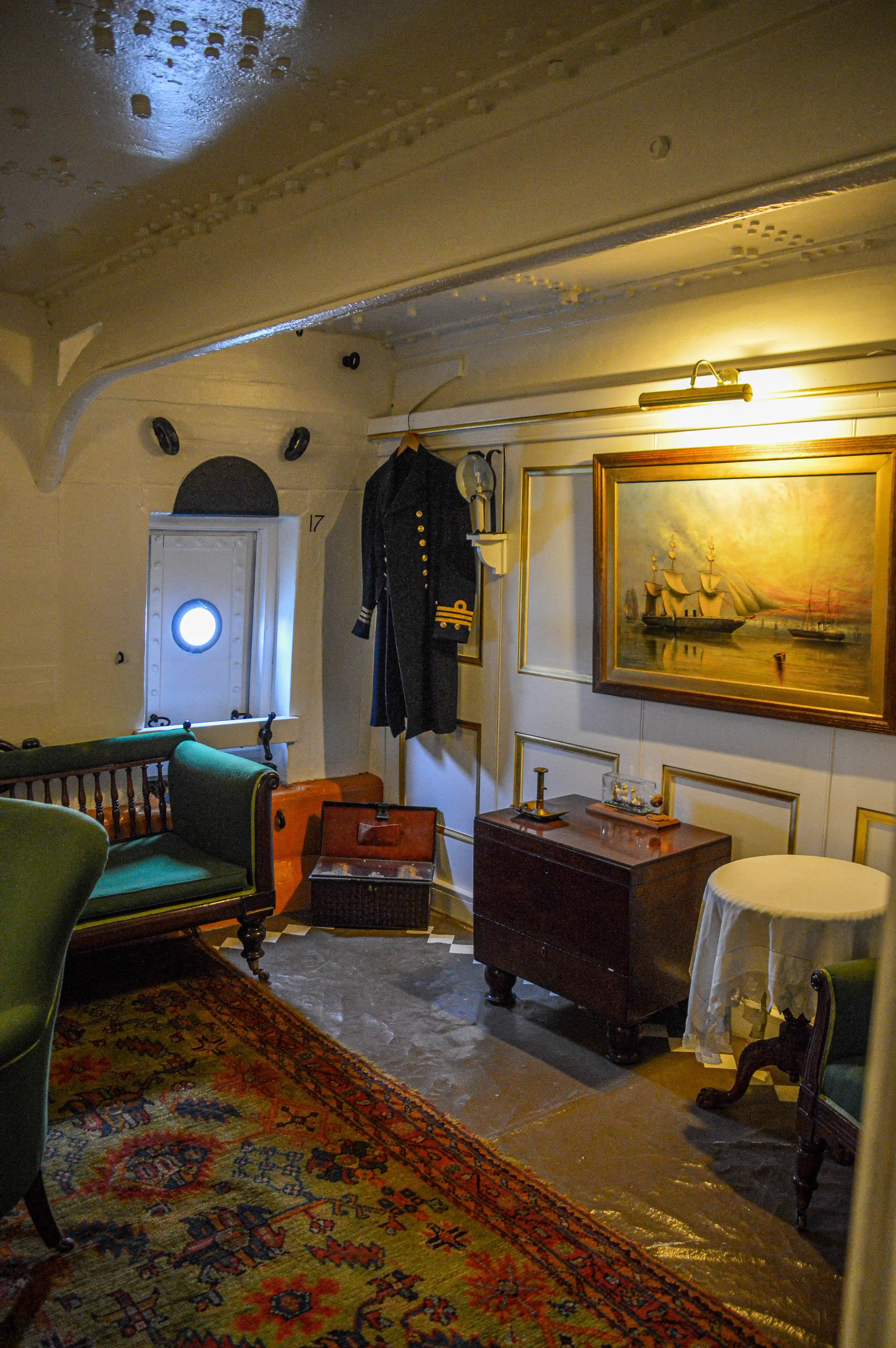 The image depicts an interior room, likely from a historical ship, featuring vintage furniture, a framed painting, a uniform hanging on a wall, and a porthole window. The room is adorned with a patterned rug and has a cozy, well-lit ambiance.