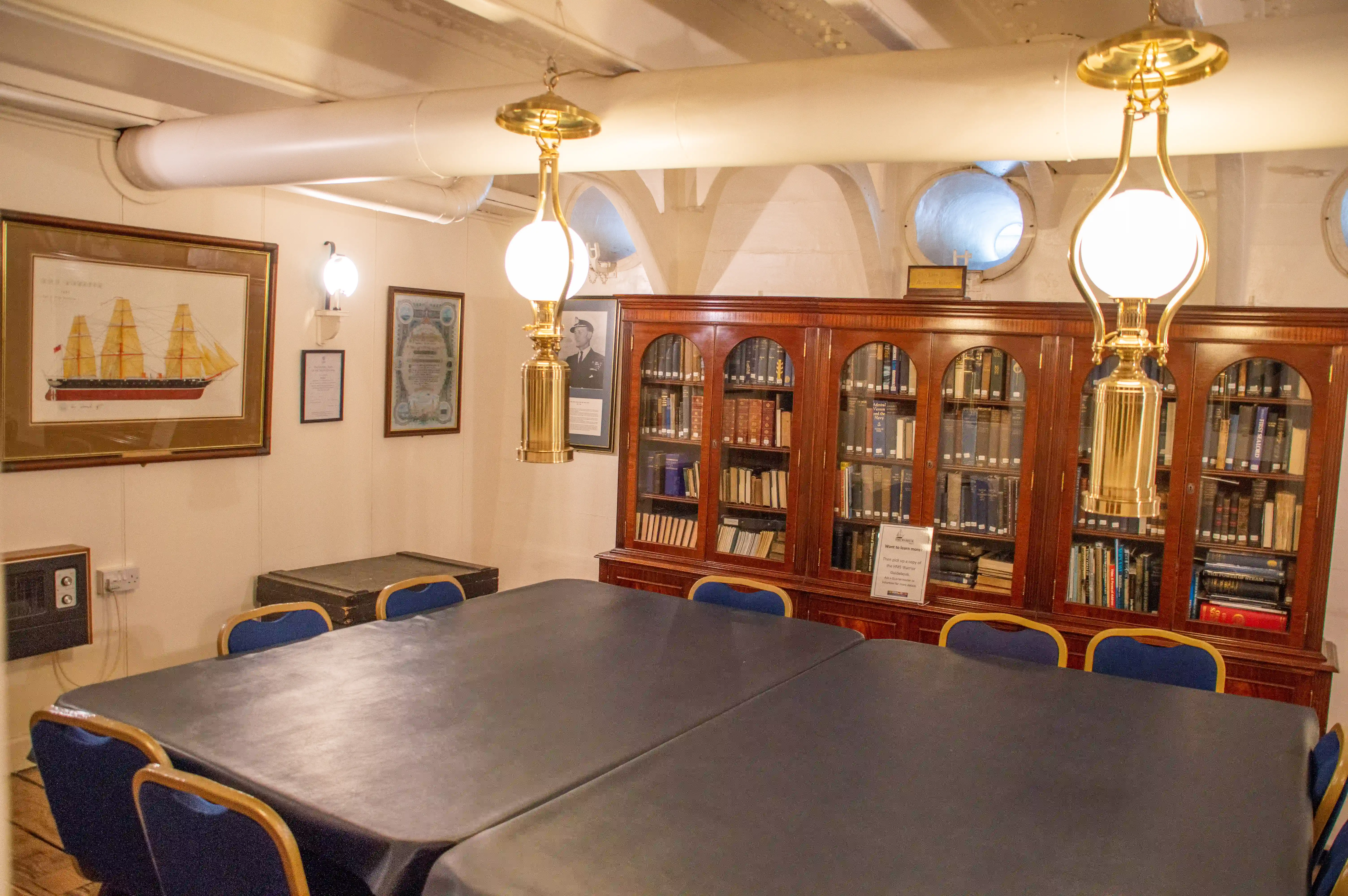The image depicts a well-appointed room that appears to be a library or a meeting room. The room features a large wooden table surrounded by blue chairs, suggesting a space for meetings or discussions. The walls are adorned with framed pictures, including a detailed drawing of a ship and a photograph of a person in uniform. The room is illuminated by elegant brass lamps hanging from the ceiling. A large wooden bookshelf filled with books and a sign indicating a 'Silent Room' further emphasize the room's purpose as a quiet space for reading or study.