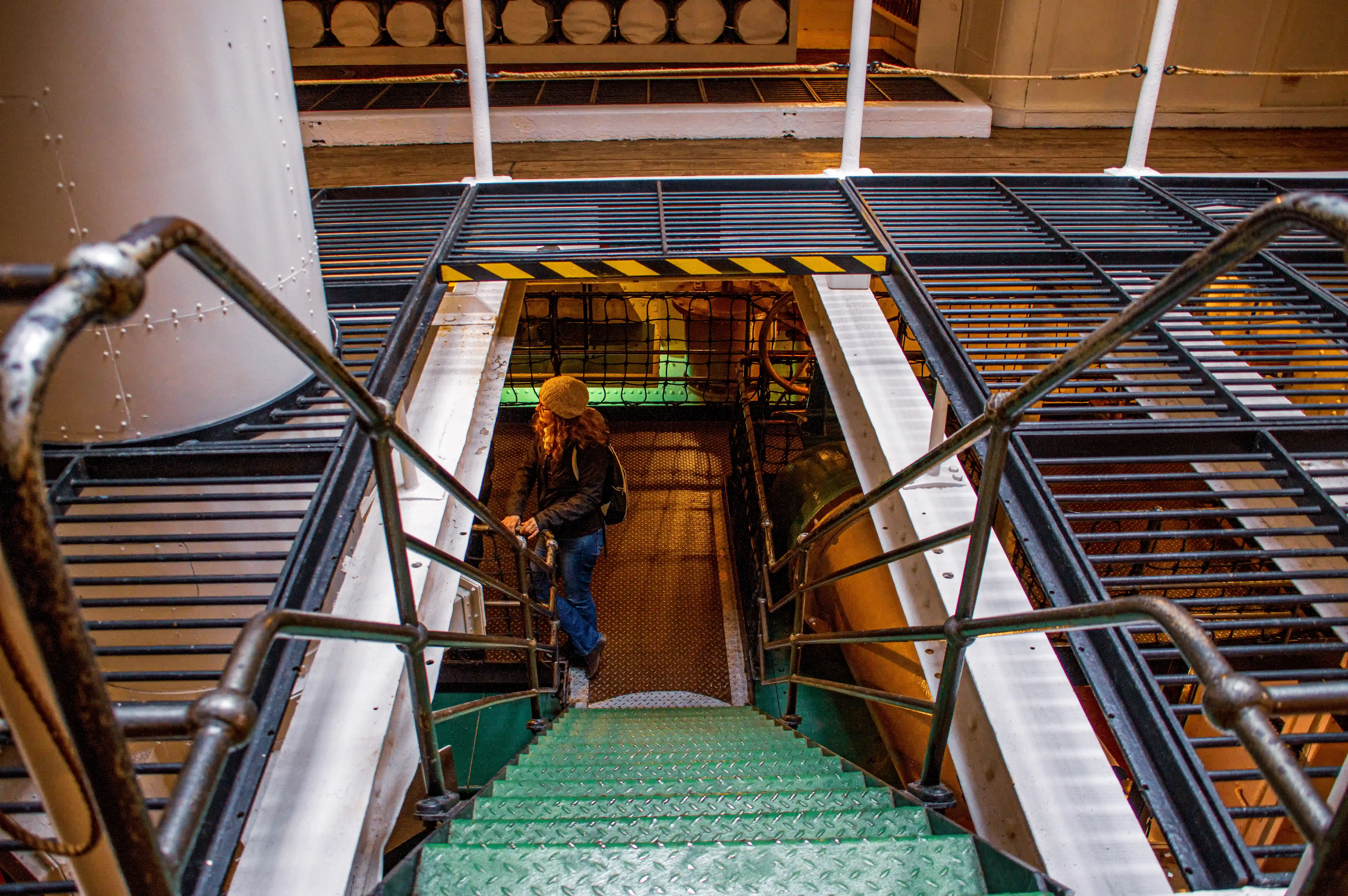 The image shows a steep, narrow ladder leading upwards within an enclosed space. The ladder is secured with ropes on either side, and the area appears to be part of an industrial or maritime setting, possibly a ship, given the visible structure and stored items like barrels and other equipment.
