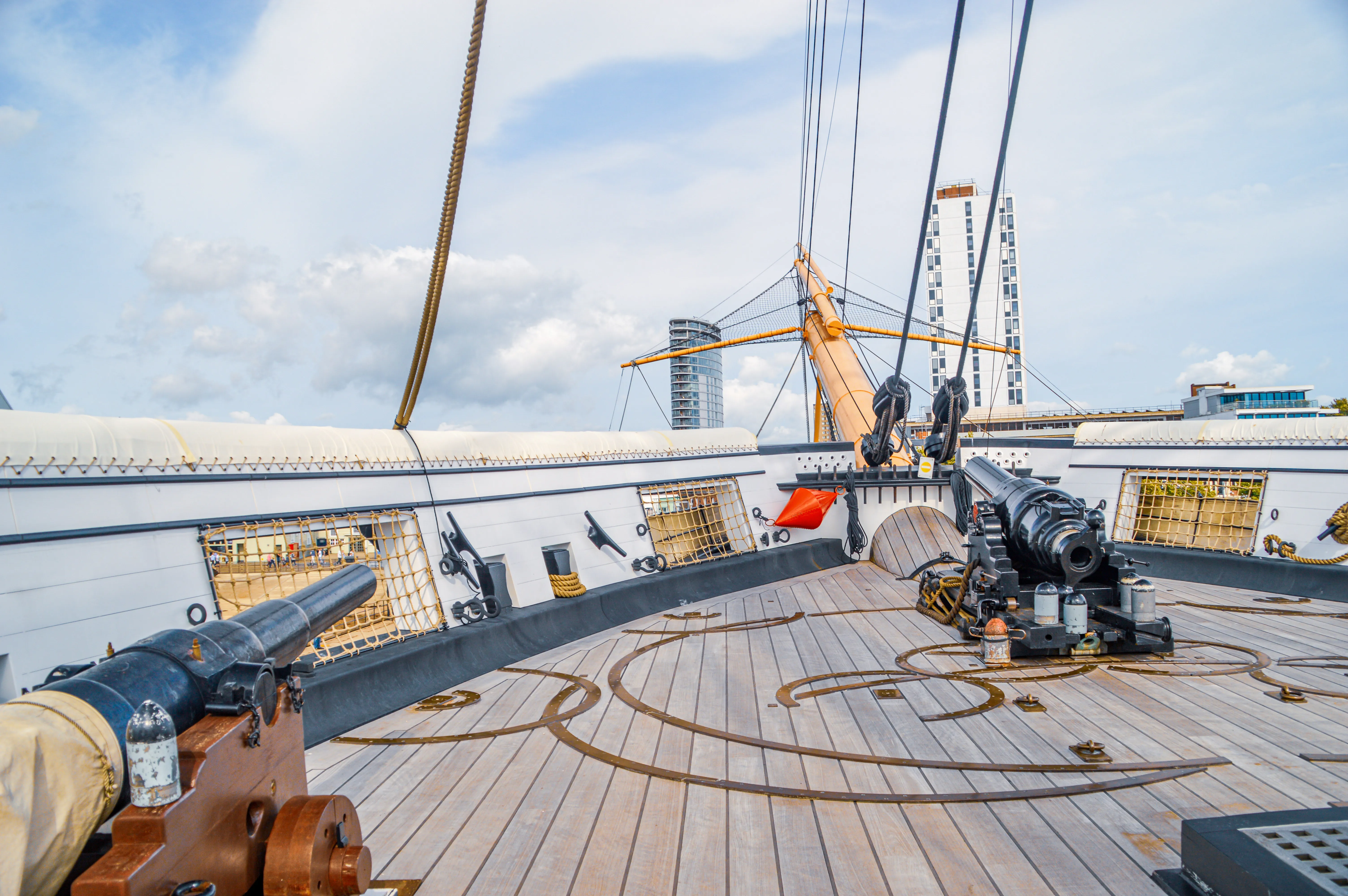 This image offers a fascinating glimpse onto the wooden deck of what appears to be a historic sailing ship. Two cannons are prominently positioned, one in the foreground on the left and another on the right. The deck itself is laid with wooden planks and features interesting circular metal inlays. In the background, modern buildings and a ship's mast create a striking contrast between old and new. The sky above is partly cloudy, suggesting a pleasant day for exploring.